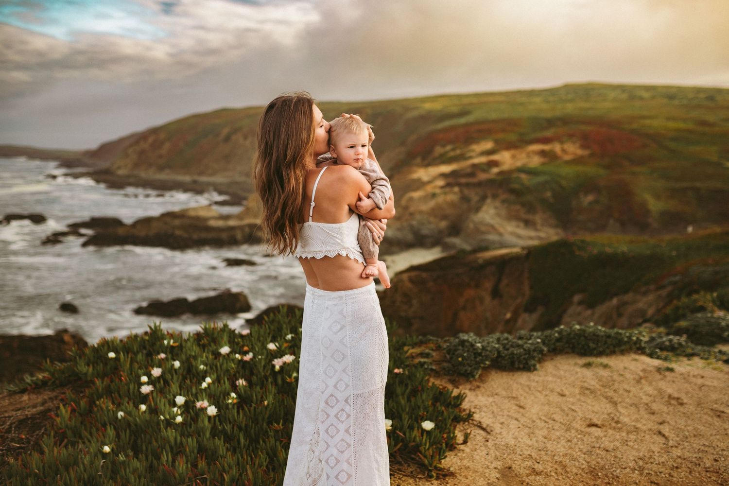 A mother stands atop a cliff overlook and holds her son in her arms while the sun is setting and the ocean waves crash below.