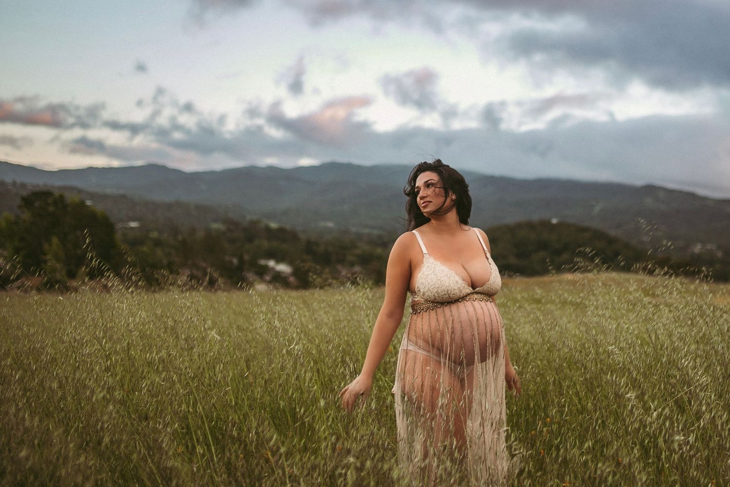 A pregnant woman stands in a tall grassy field wearing a lace skirt and looking towards the mountains and sunset.