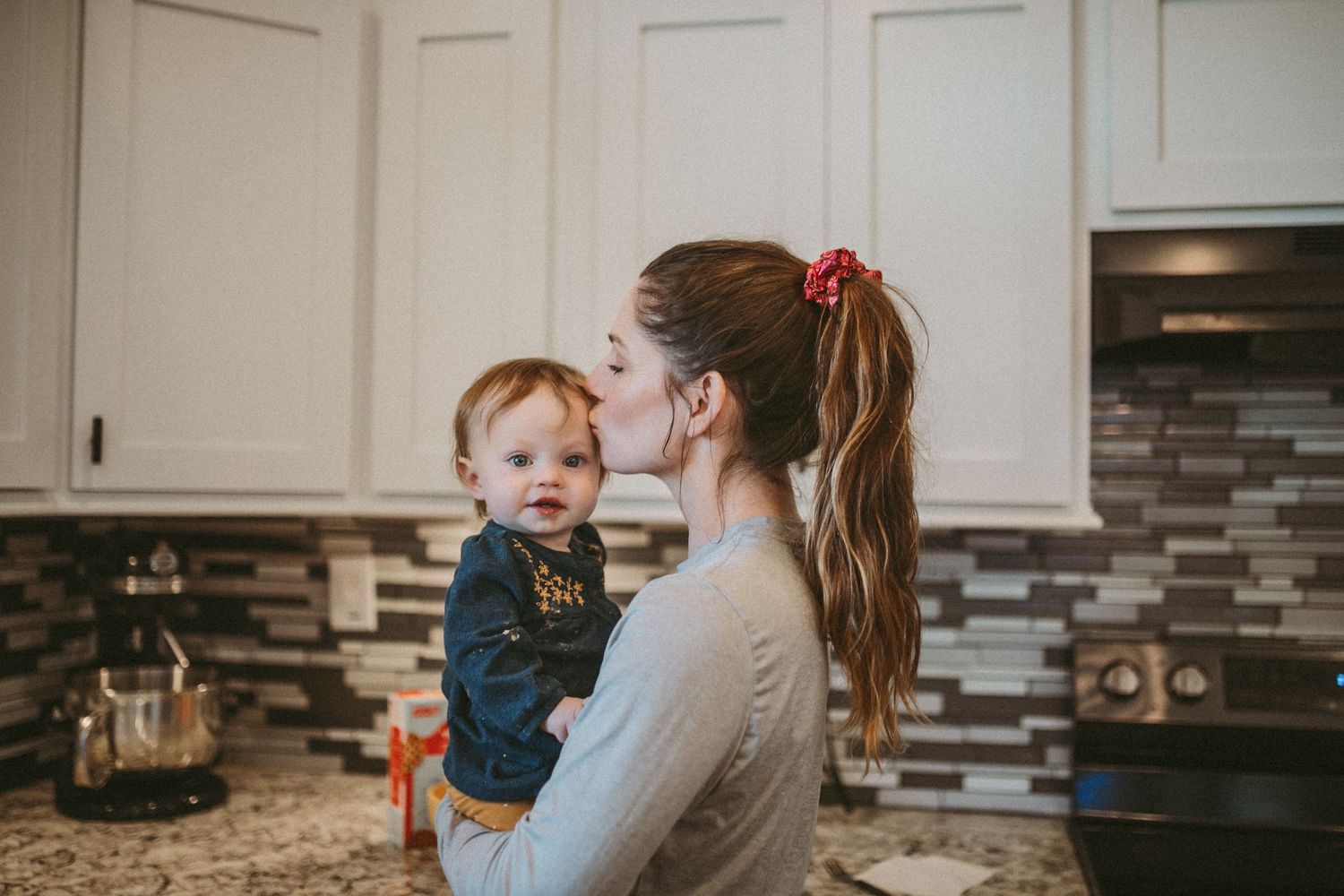 A mother kisses her young daughter on the temple while standing in the kitchen, photographed by Kristy Baker Photography.