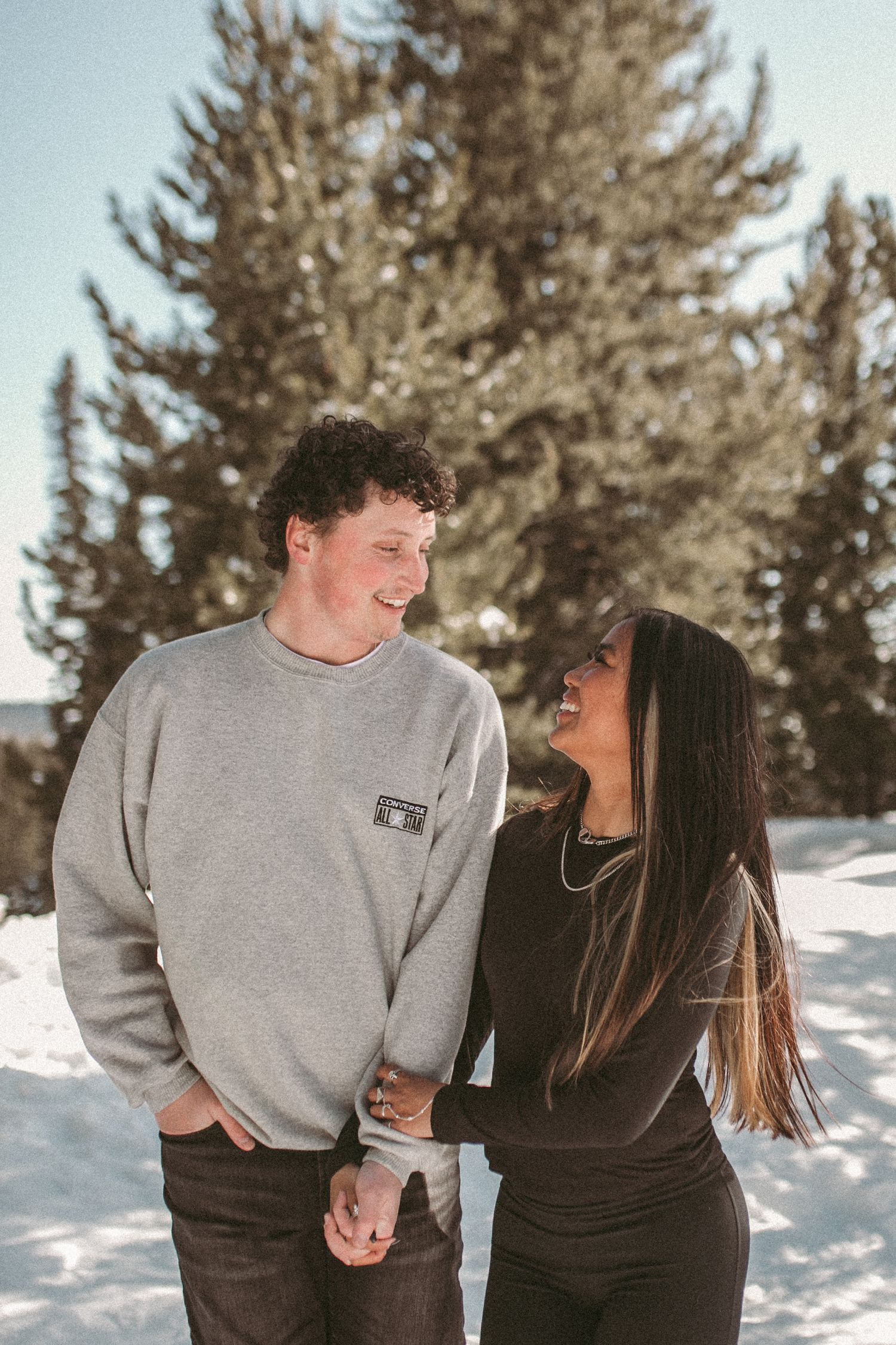A happy couple smiling and walking by pine trees in the winter, photographed by Kristy Baker Photography in Steamboat Springs, Colorado.