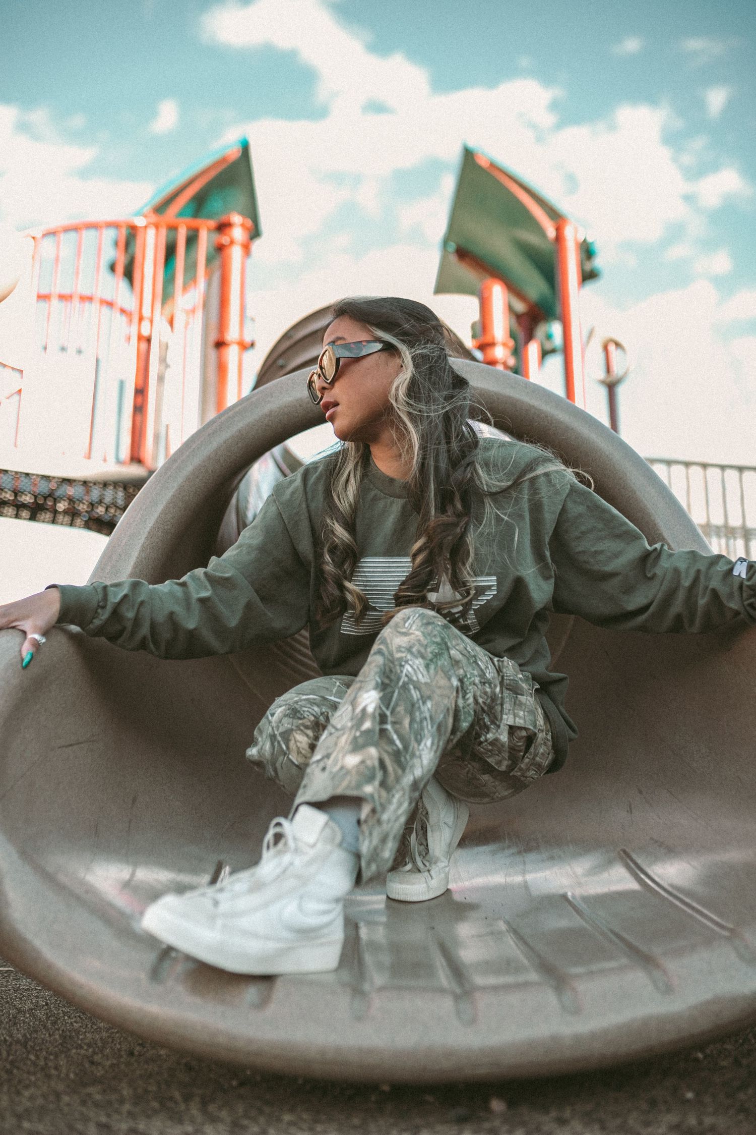 A woman squats on the end of a slide and looks outwardly while wearing sunglasses.