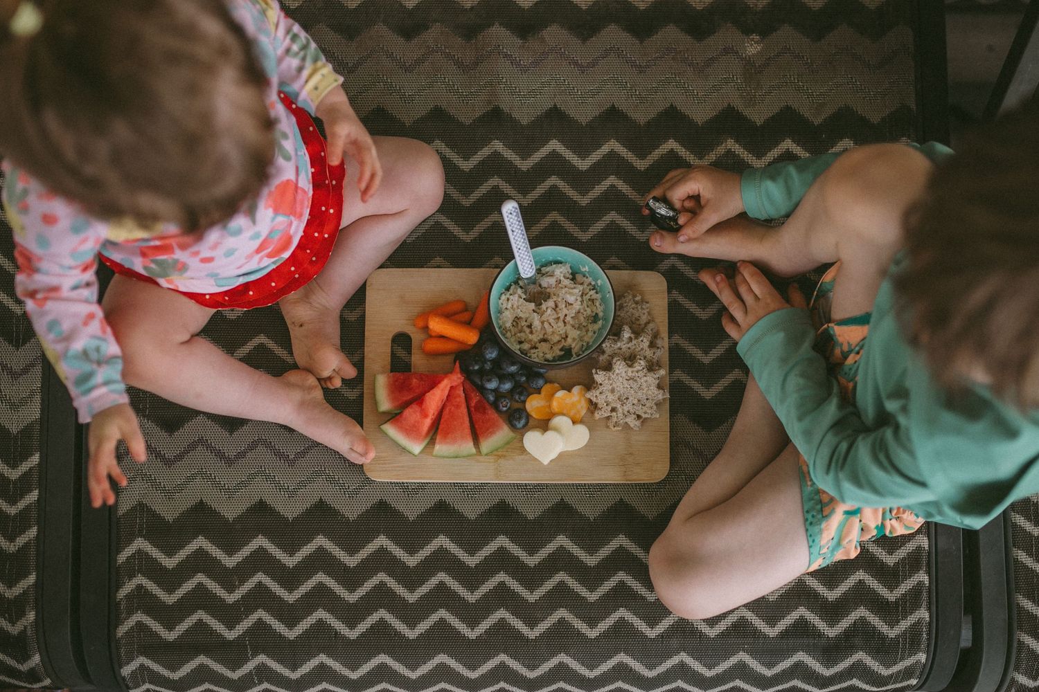 A young boy and girl are sitting and about to eat a kid-approved charcuterie board with watermelon, blueberries, cheese, and tuna sandwich.