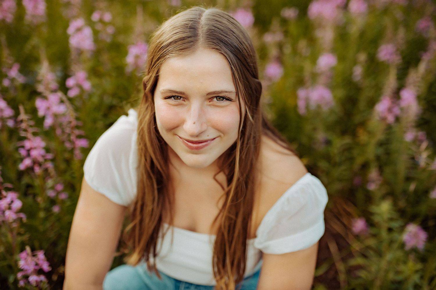 A high school senior girl sits amidst a field of purple fireweed wildflowers and grins as she looks at the camera.