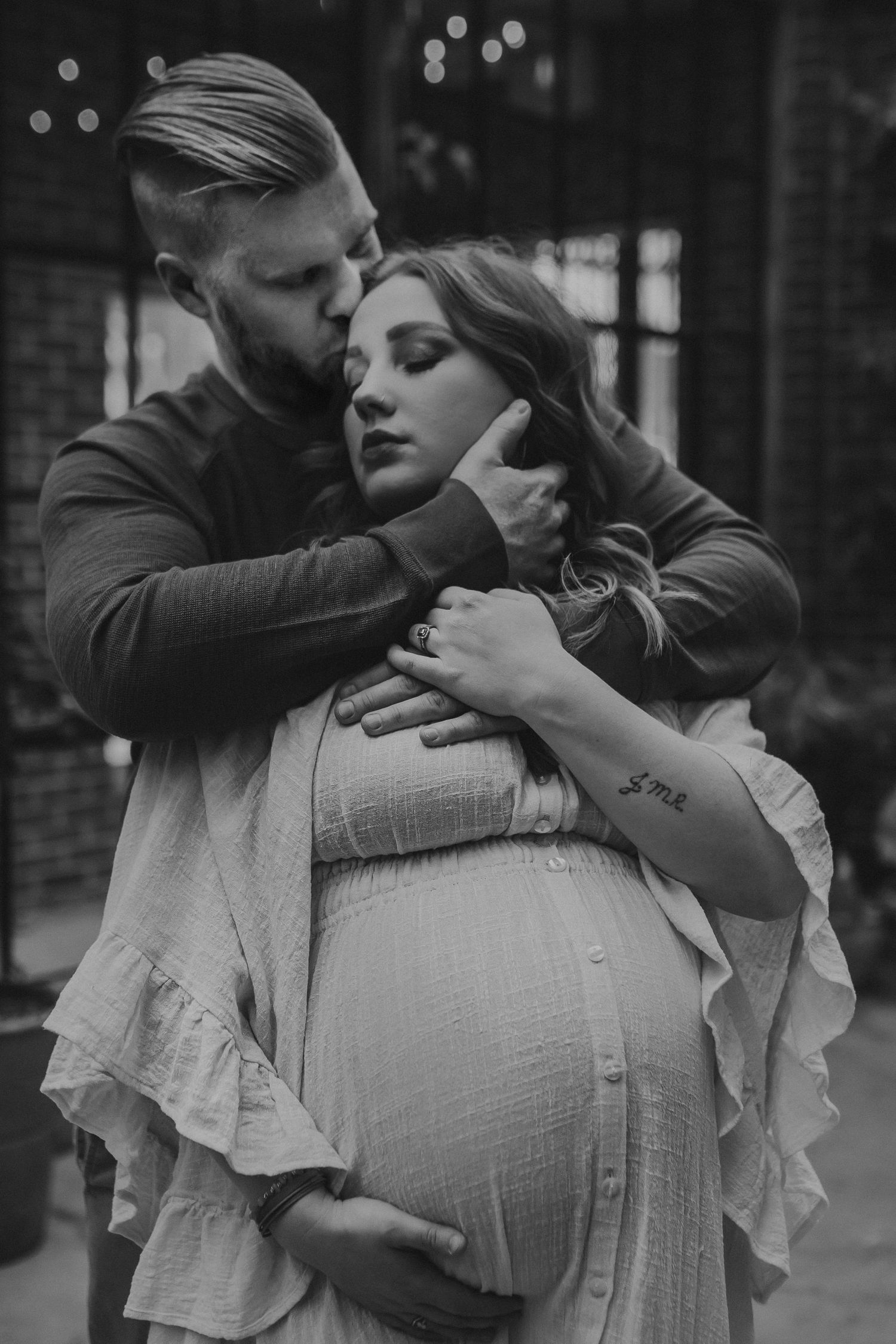 A pregnant mother is embraced by her husband from behind as he kisses her on her temple, photographed by Kristy Baker Photography.
