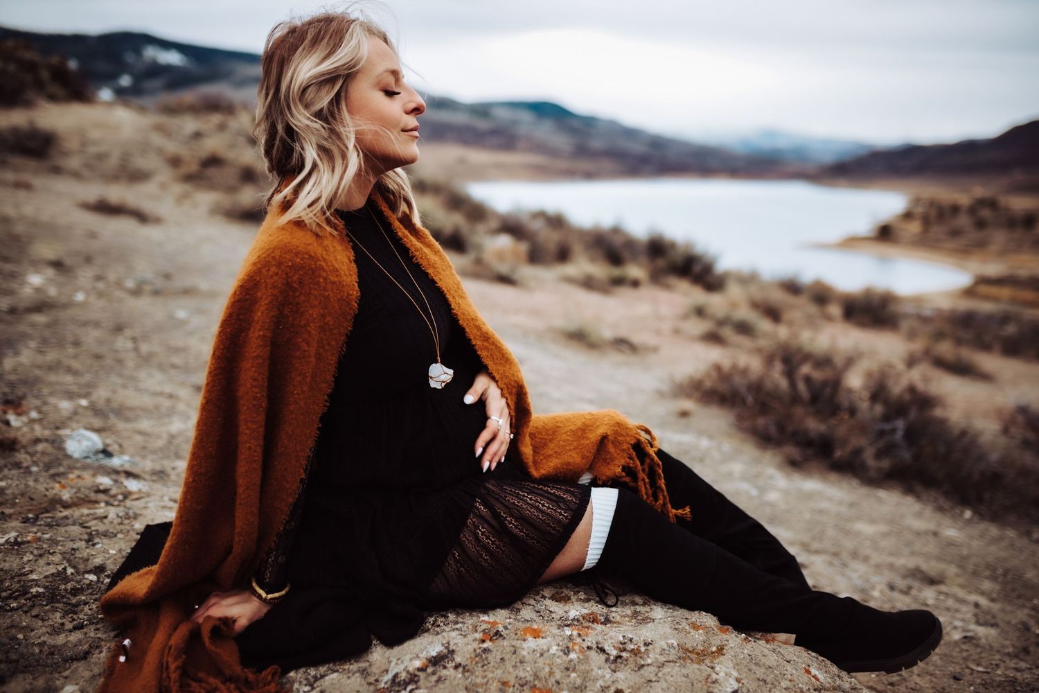 A pregnant woman in a black dress sitting and cradling her belly, photographed by Kristy Baker Photography in Steamboat Springs, Colorado.
