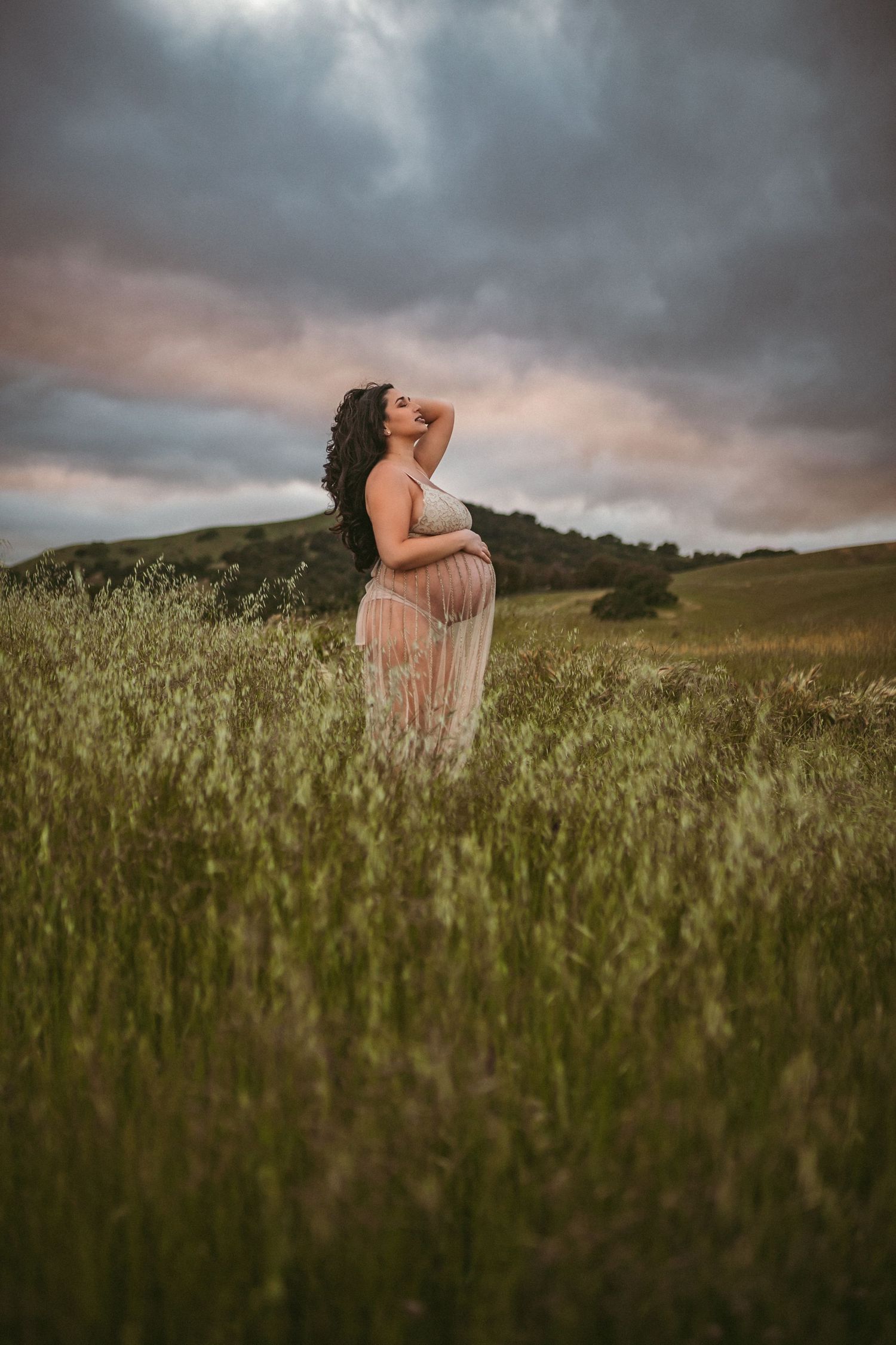 A pregnant woman standing in tall grass cradles her belly while looking at the sunset, photographed by Kristy Baker Photography.