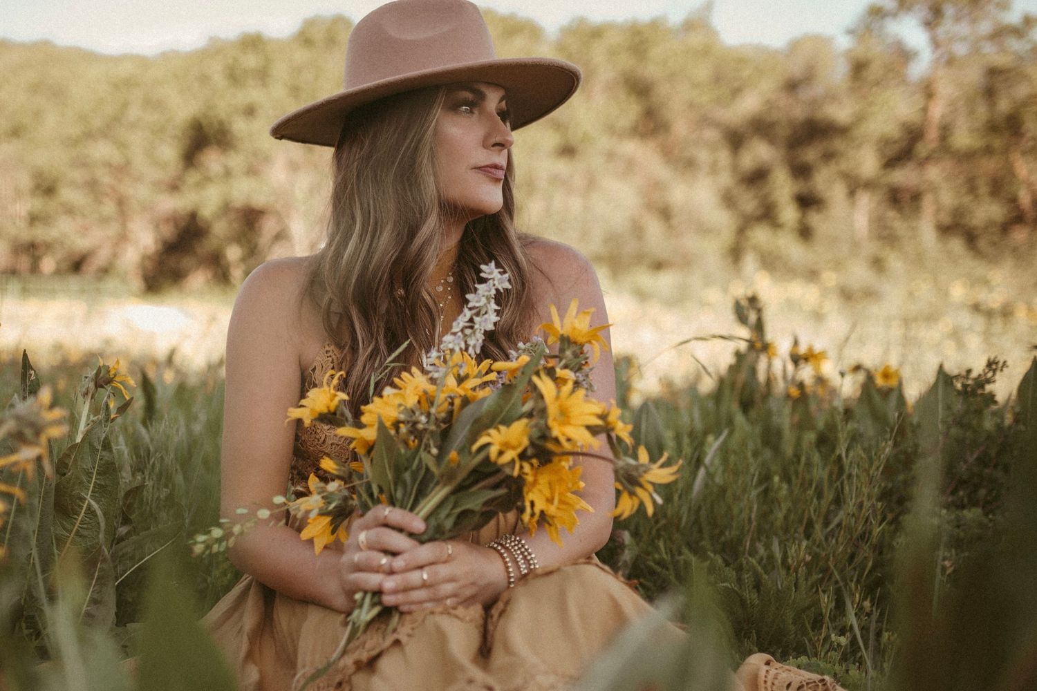 A woman wearing a brown hat looks off into the distance as she sits in a field and holds a bouquet of yellow and purple wildflowers.