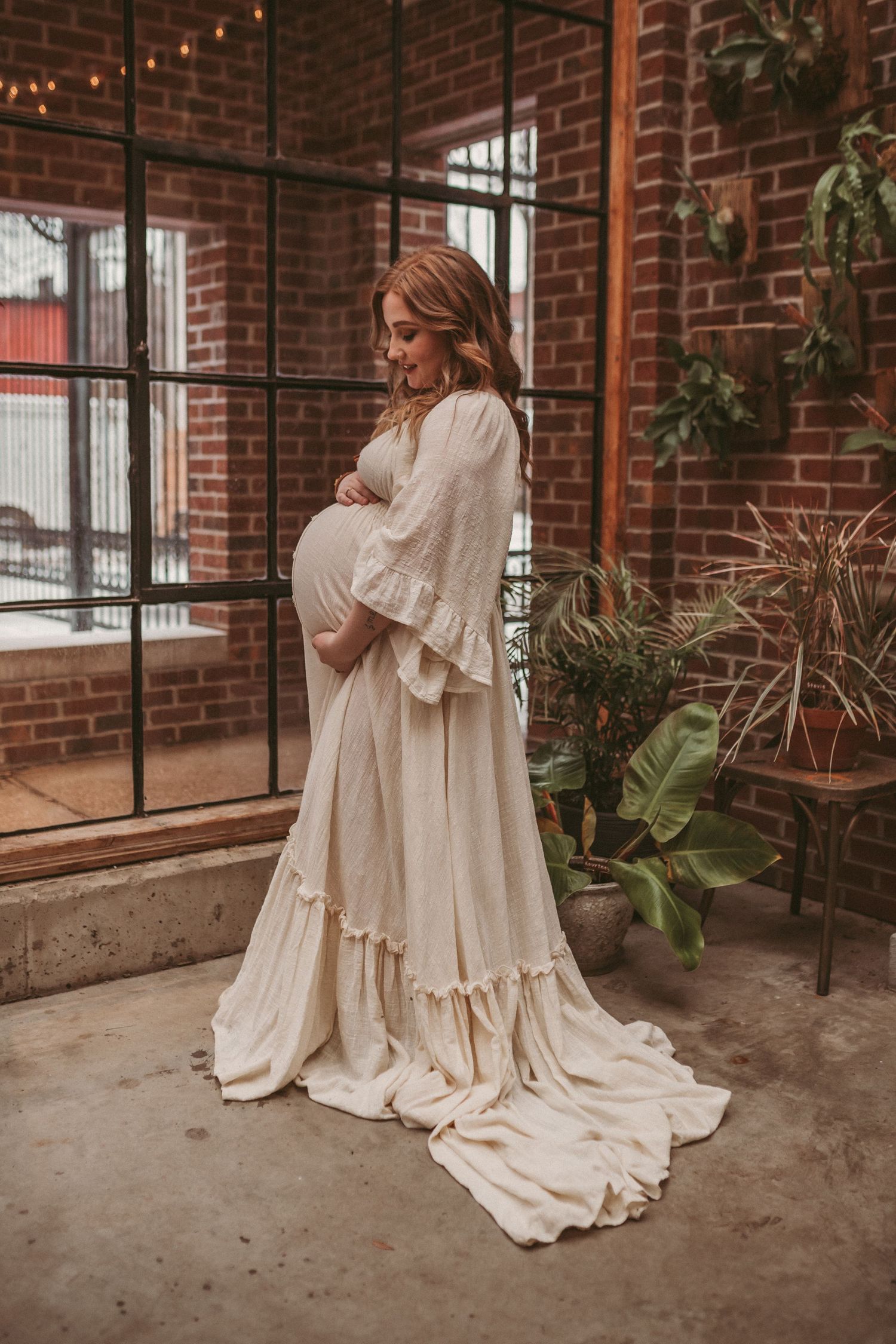 A pregnant mother stands inside a brick building looking down at her belly, photographed by Kristy Baker Photography.