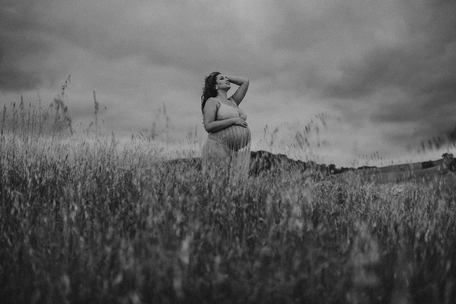 A pregnant woman standing in tall grass cradles her belly while looking at the sunset, photographed by Kristy Baker Photography.