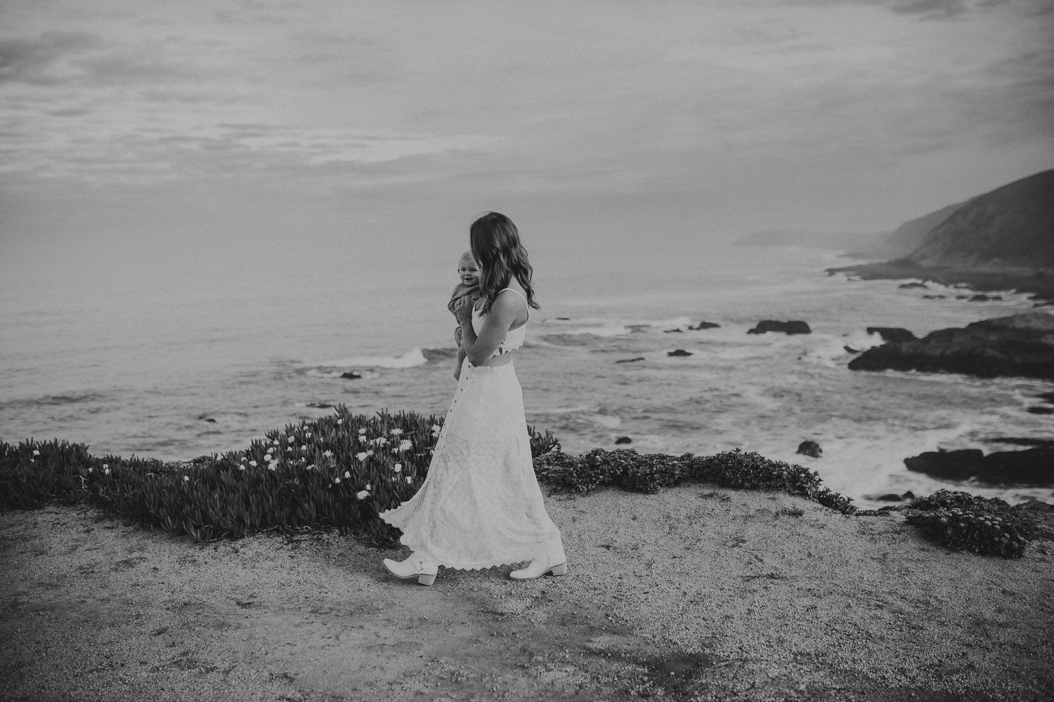 A mother holding her little boy is walking on a mountain while the ocean water down below crashes amongst the shore.