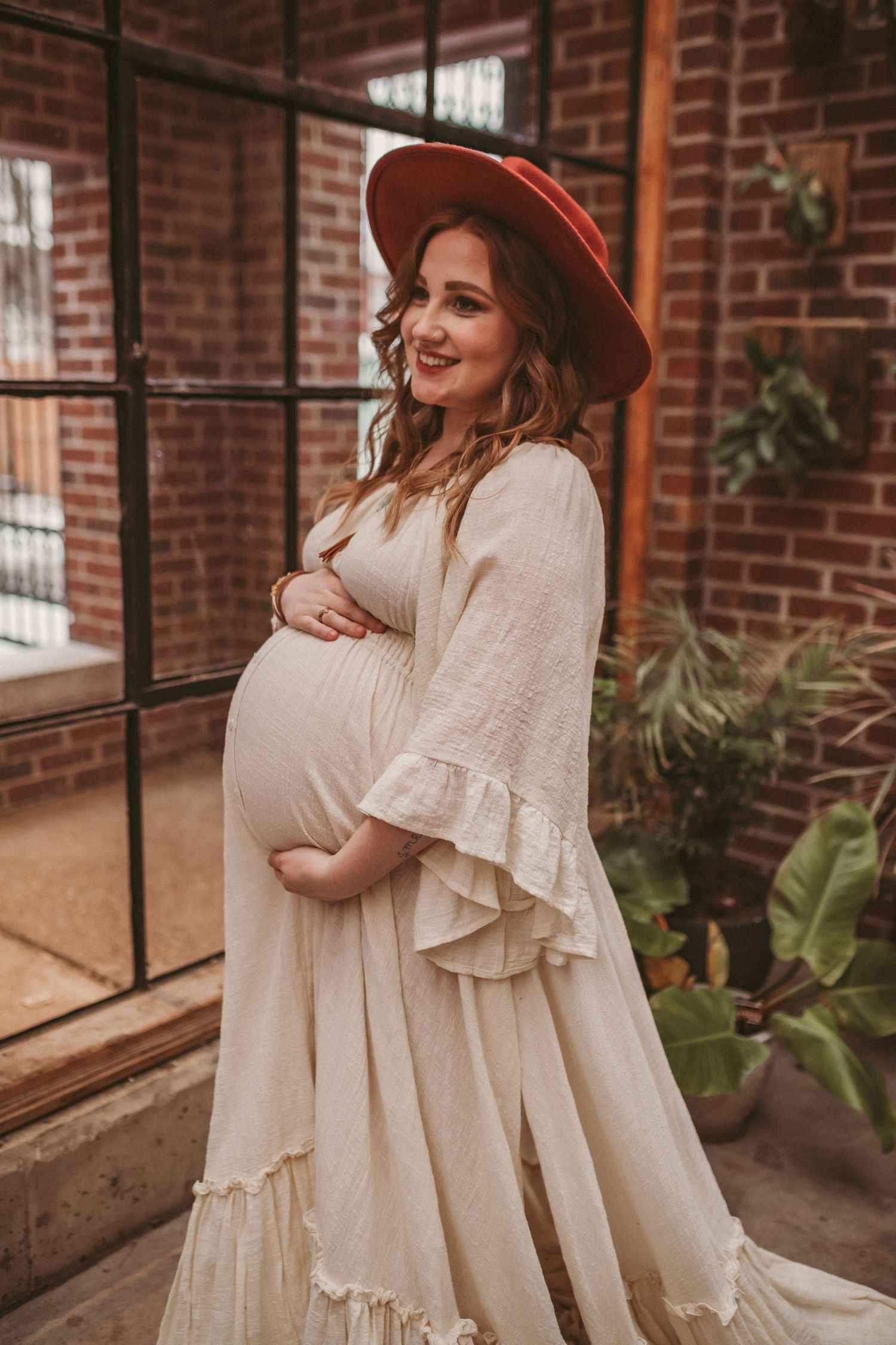 A pregnant mother stands inside a brick building looking happy about her growing belly, photographed by Kristy Baker Photography.