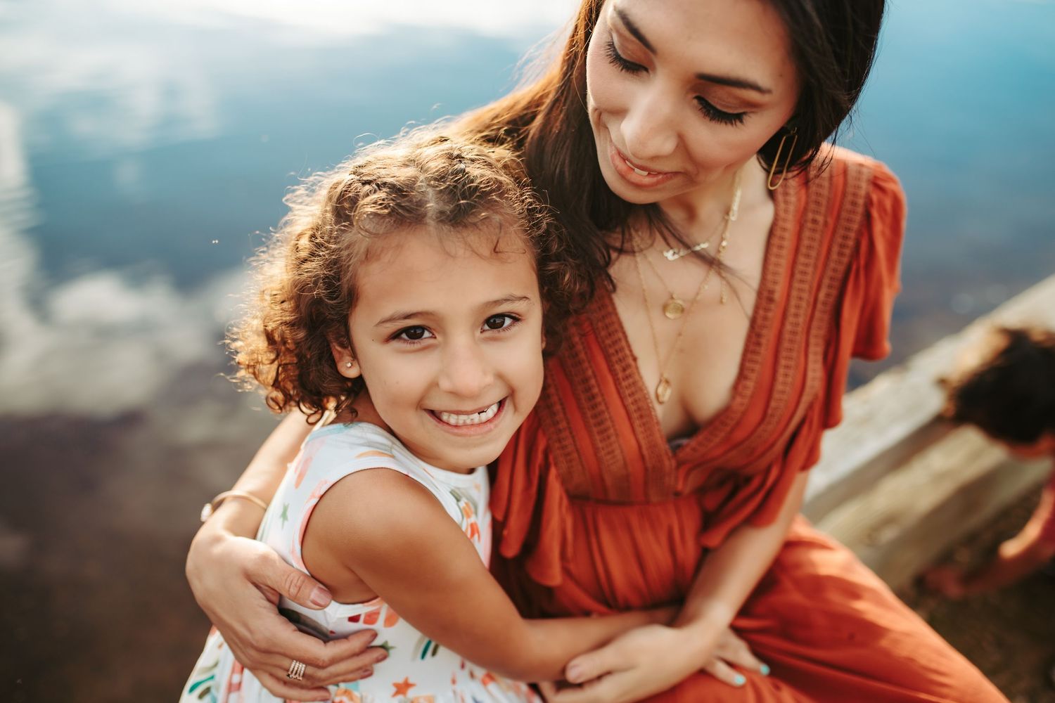 A mother sits next to her young daughter near a lake and embraces her from the side, photographed by Kristy Baker Photography.