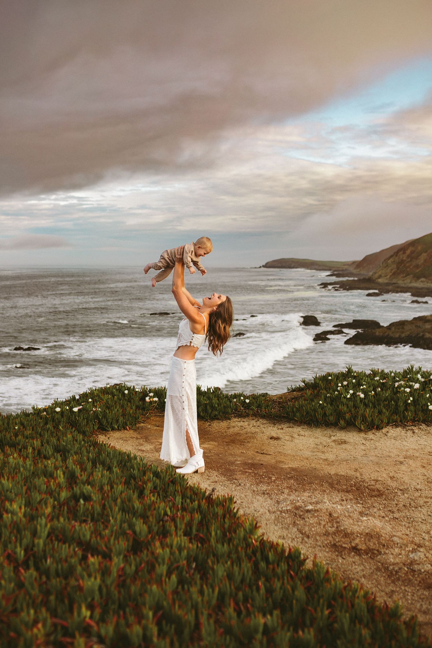 A mother is standing atop a cliff and lifts her baby boy high in the air and looks at him smiling, photographed by Kristy Baker Photography.