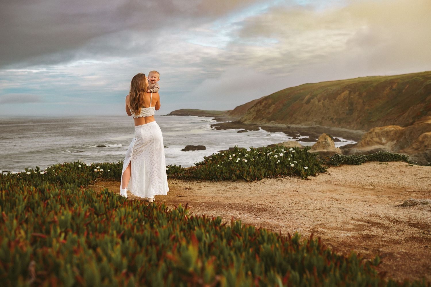 A mother stands atop a cliff overlook and holds her son in her arms while the sun is setting and the ocean waves crash below.