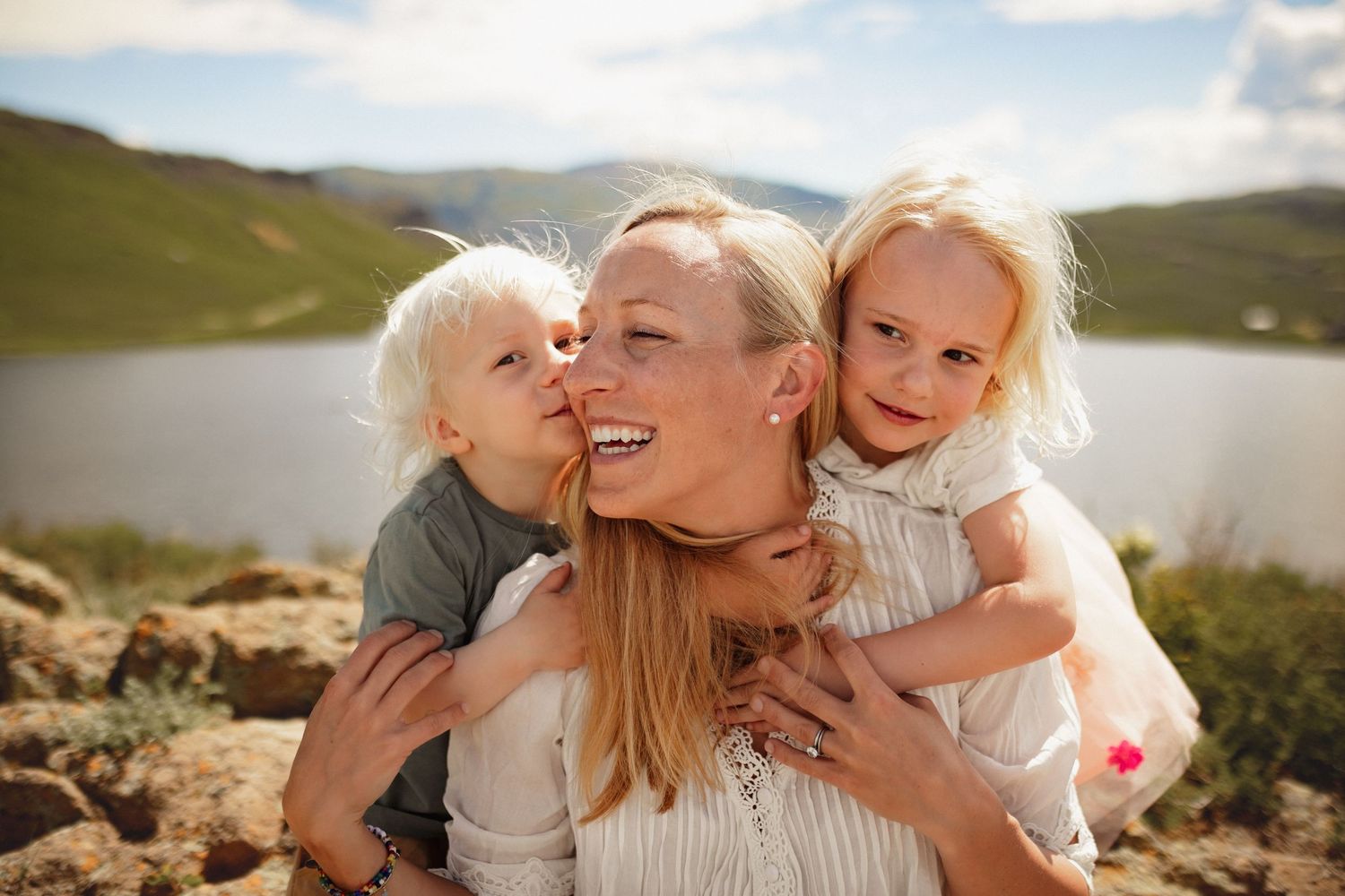 A smiling mother is embraced by her two young children while on top of a mountain with a lake in the distance, photographed by Kristy Baker.