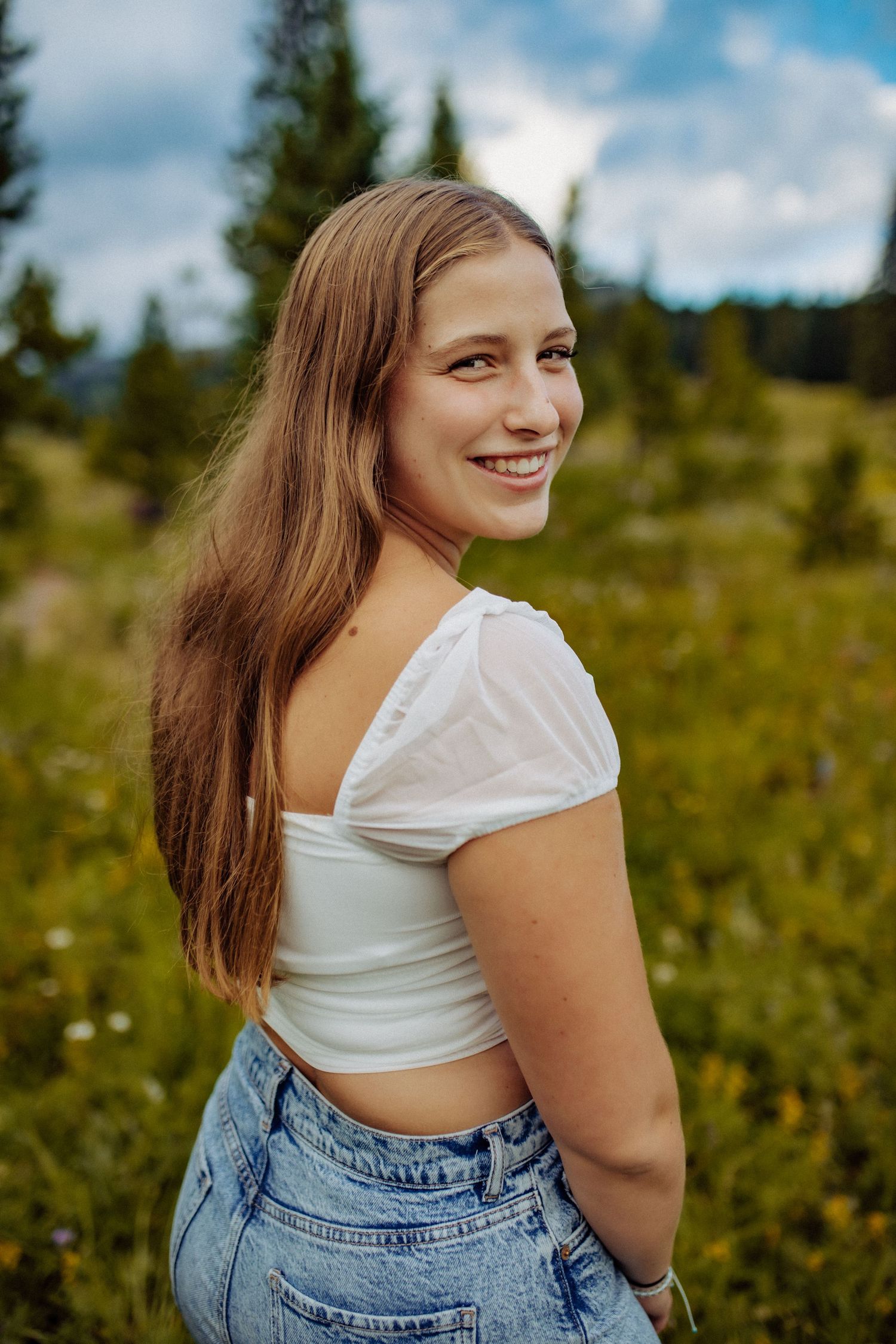 A high school senior girl wearing a white shirts and jeans is smiling as she peers over her shoulder.