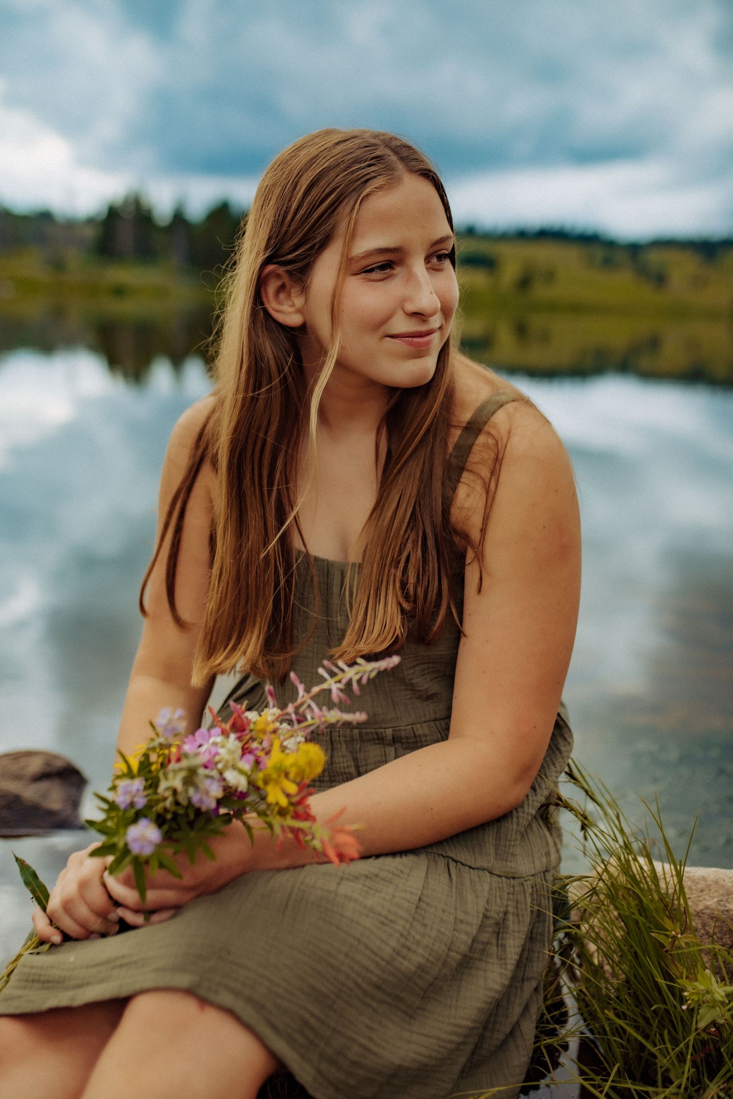 A high school senior girl sits upon a log near a lake and holds a bouquet of wildflowers as she looks towards the clouds.