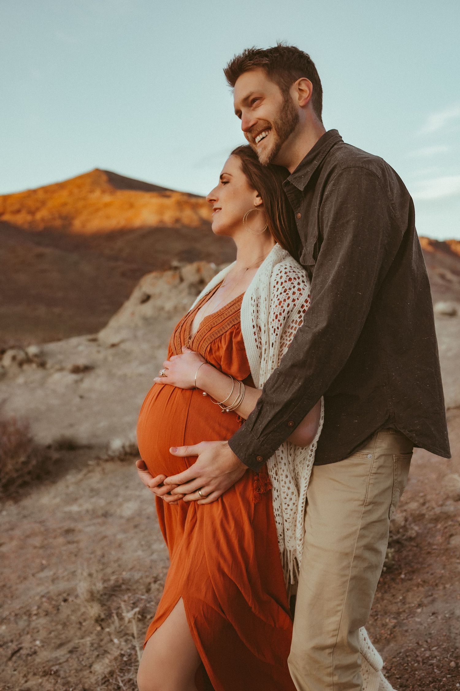 A pregnant mother cradling her belly with her husband standing behind her, photographed by Kristy Baker Photography.