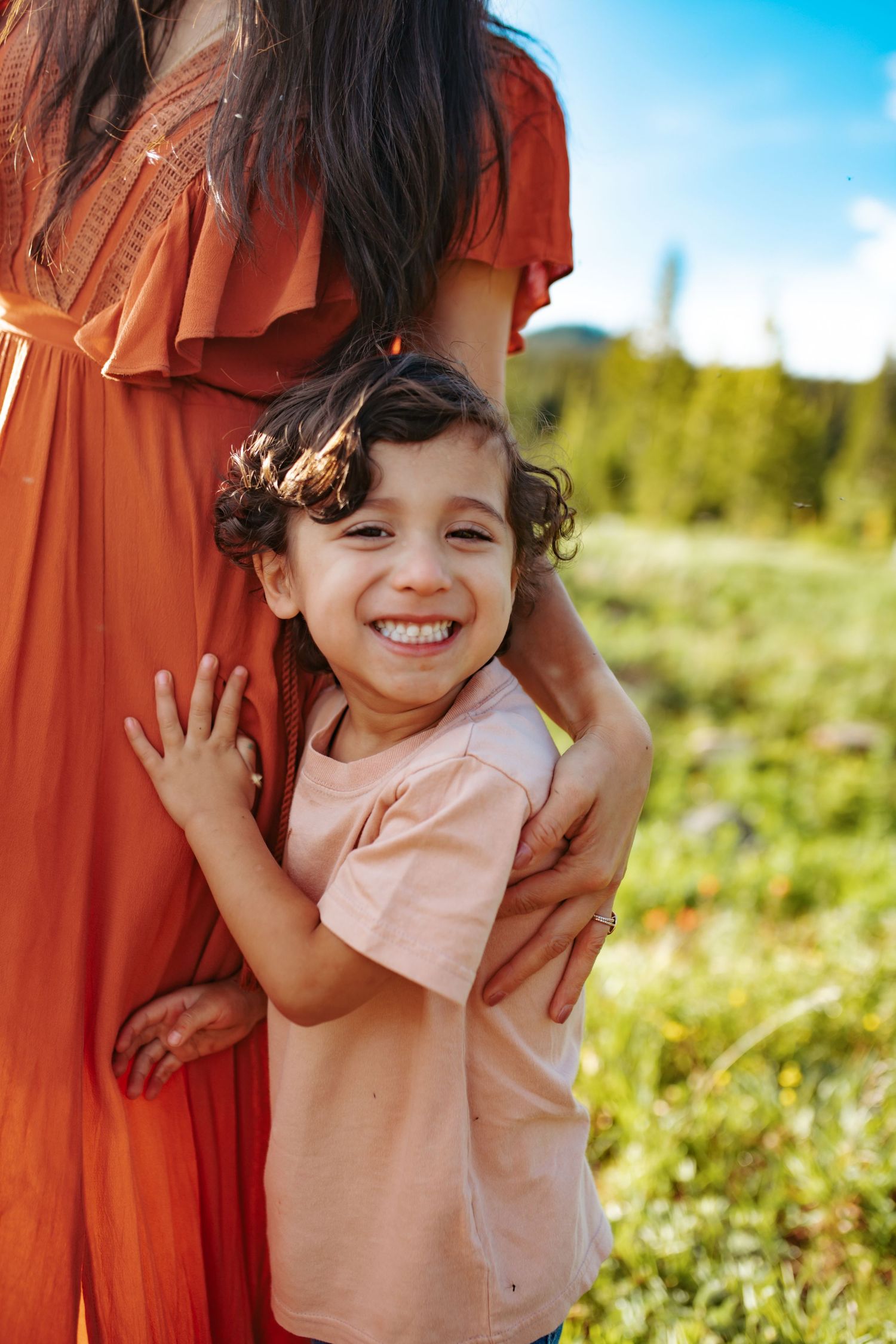 A young boy is smiling big and hugging his mother's leg, photographed by Kristy Baker Photography.