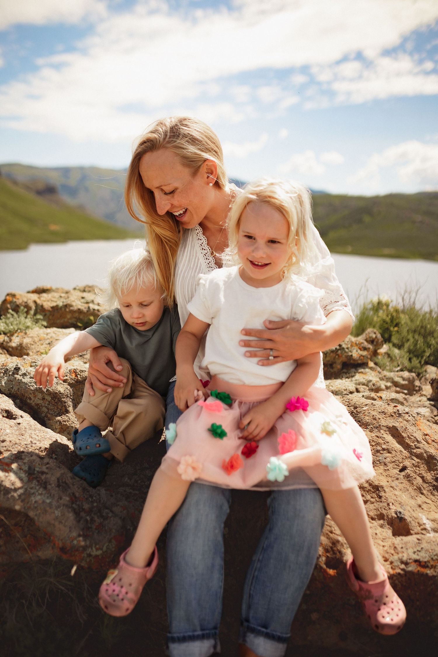 A mother sits on a rock with her daughter on her lap and her son sitting next to her, with mountains and a lake in the background.