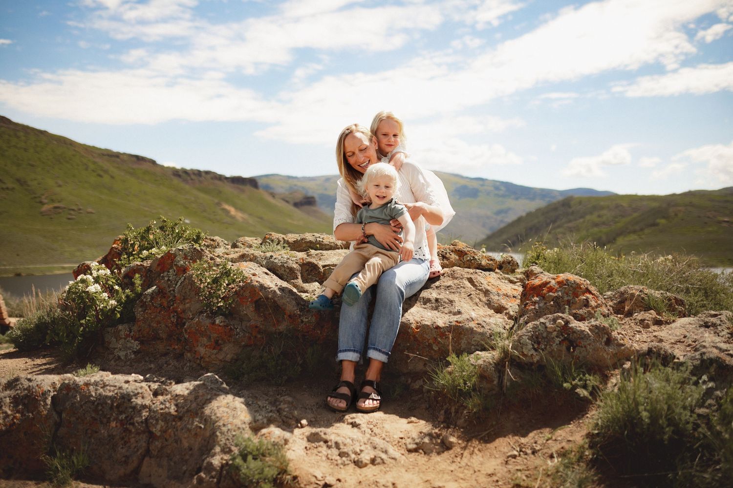 A mother sits atop a pile of rocks with her son on her lap and her daughter hugging her, photographed by Kristy Baker Photography.