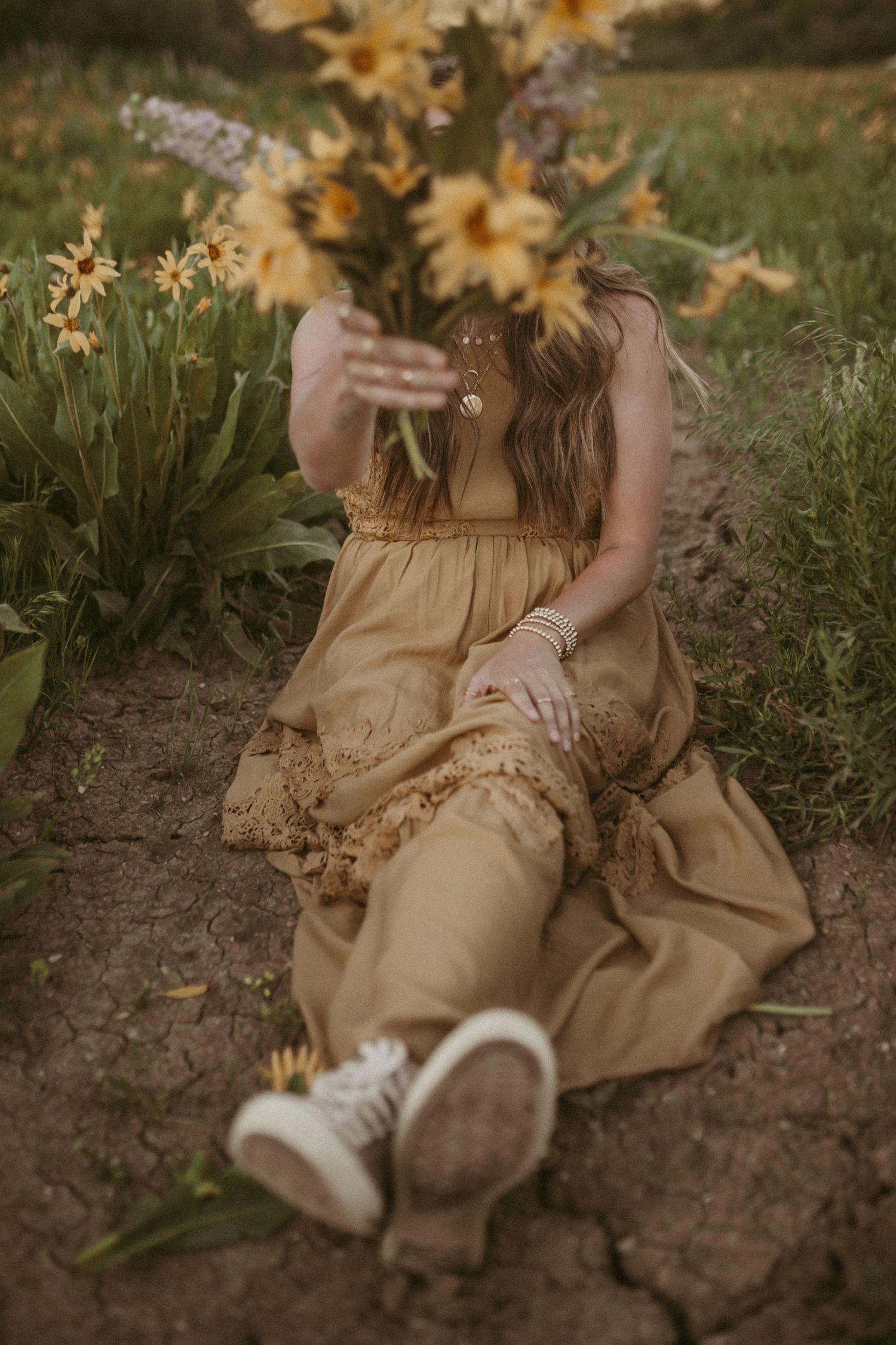 A woman sits in the dirt and holds a big bouquet of yellow wildflowers in front of her face.