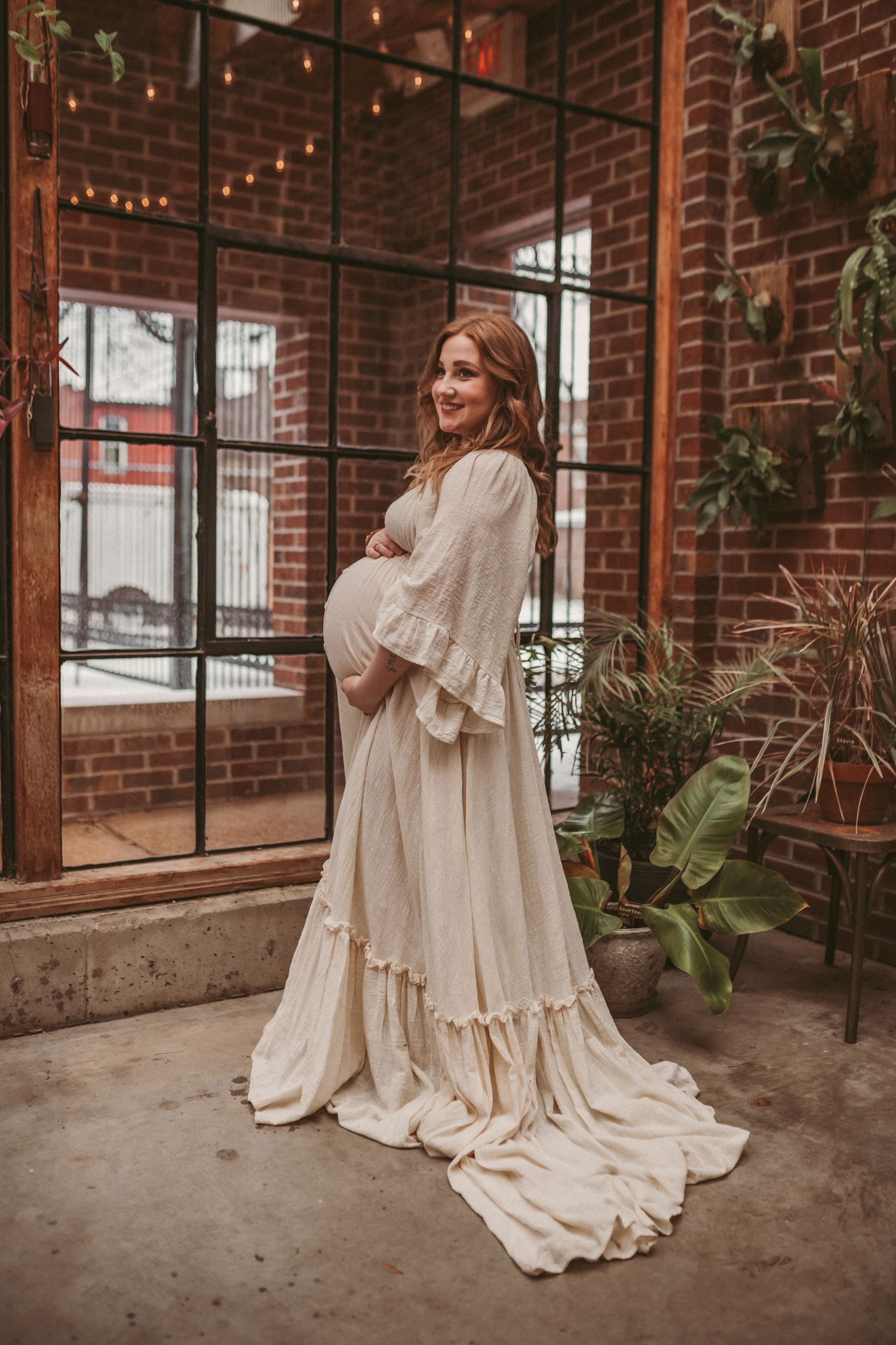 A pregnant mother stands inside a brick building looking happy about her growing belly, photographed by Kristy Baker Photography.