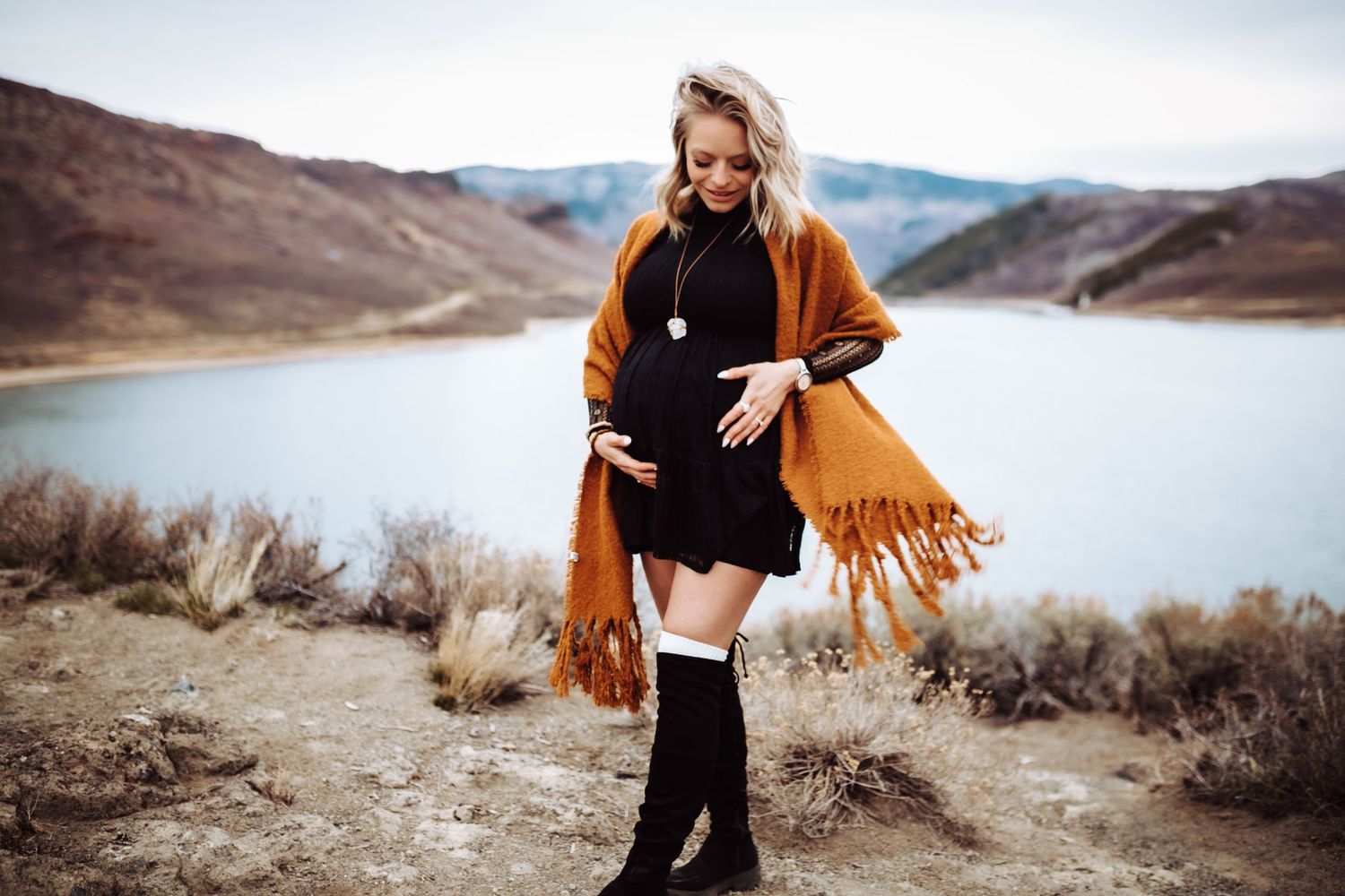 A pregnant woman wearing a black dress stands atop a hill and cradles her growing belly, photographed by Kristy Baker Photography.
