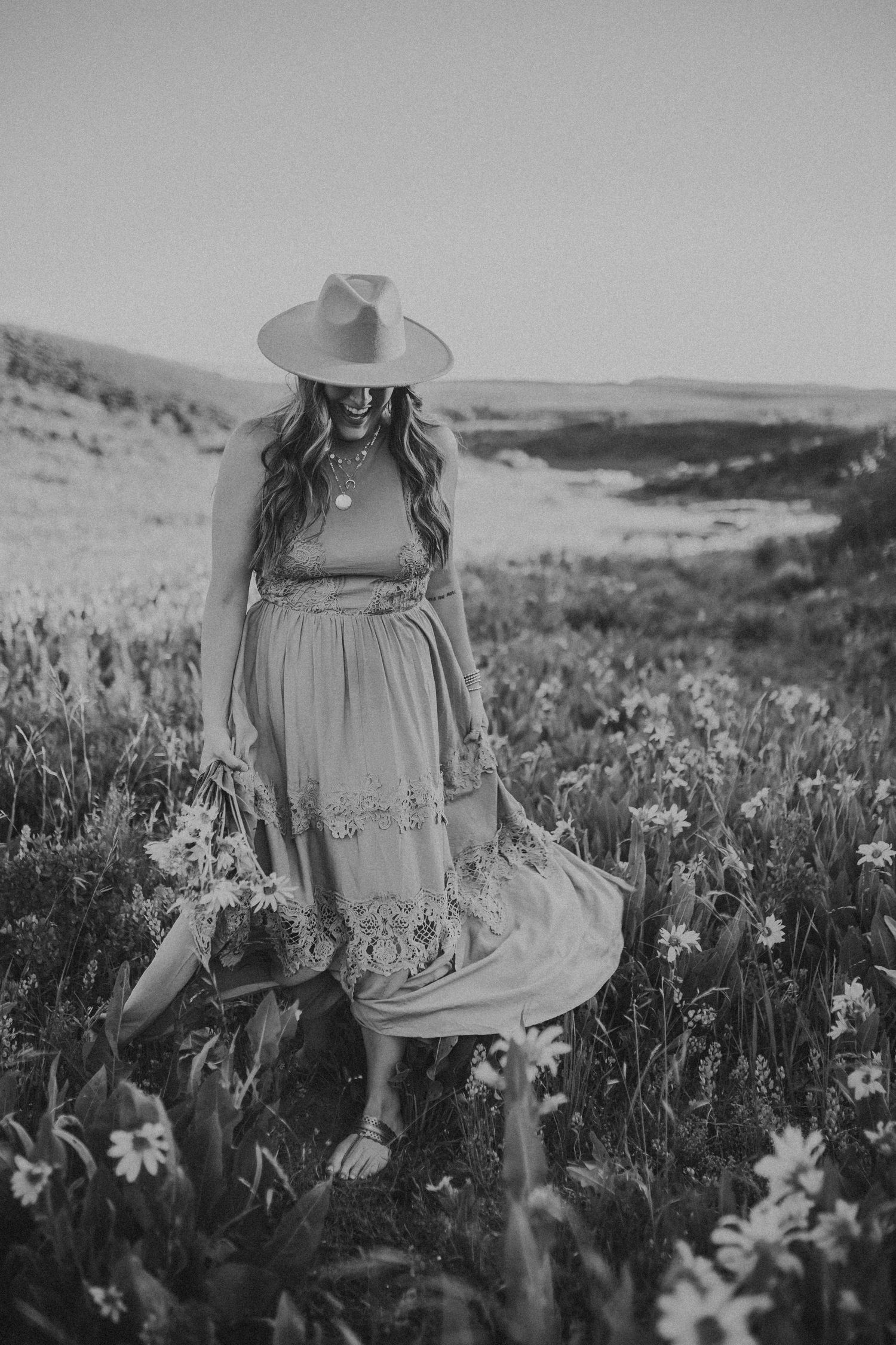 A woman in a dress and wide brimmed had looks down as she walks in a field of wildflowers while holding a bouquet.