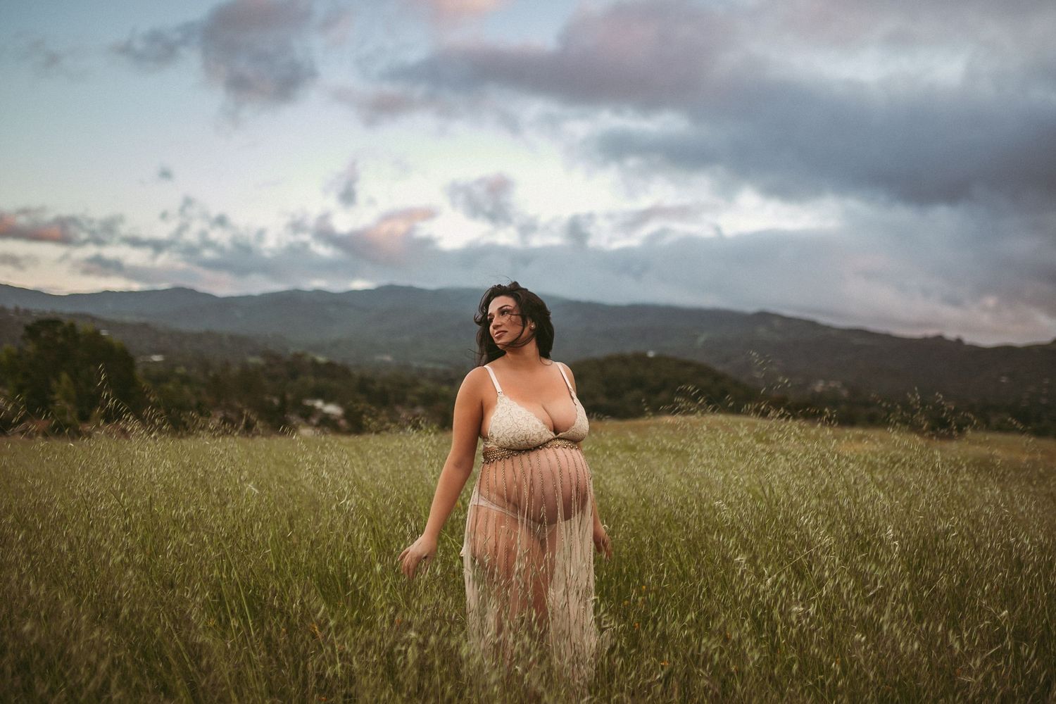 A pregnant woman standing in tall grass shows off her belly while looking at the sunset, photographed by Kristy Baker Photography.