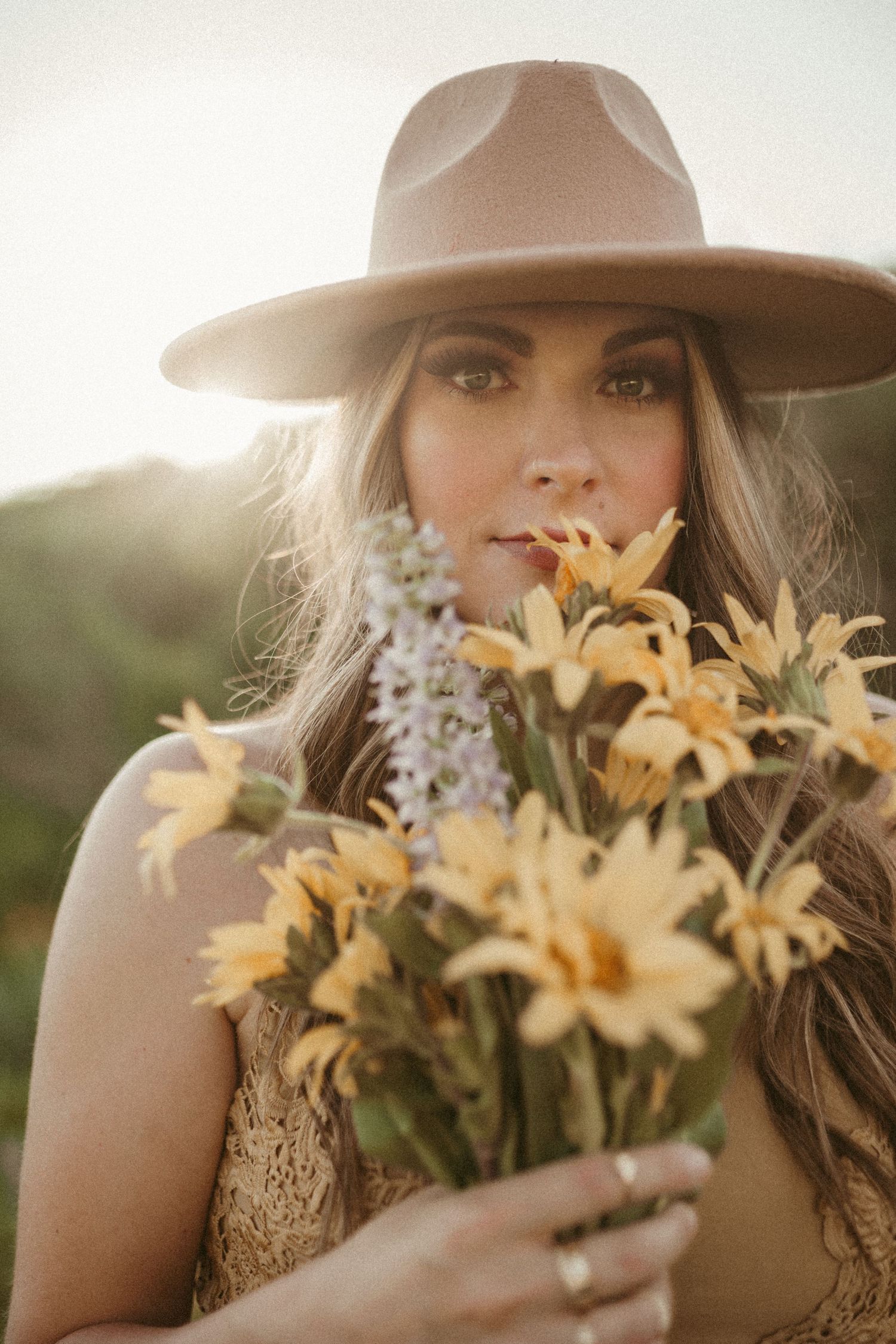 A woman wearing a light brown wide brimmed hat is holding a bouquet of yellow and purple wildflowers and looks straight into the camera.