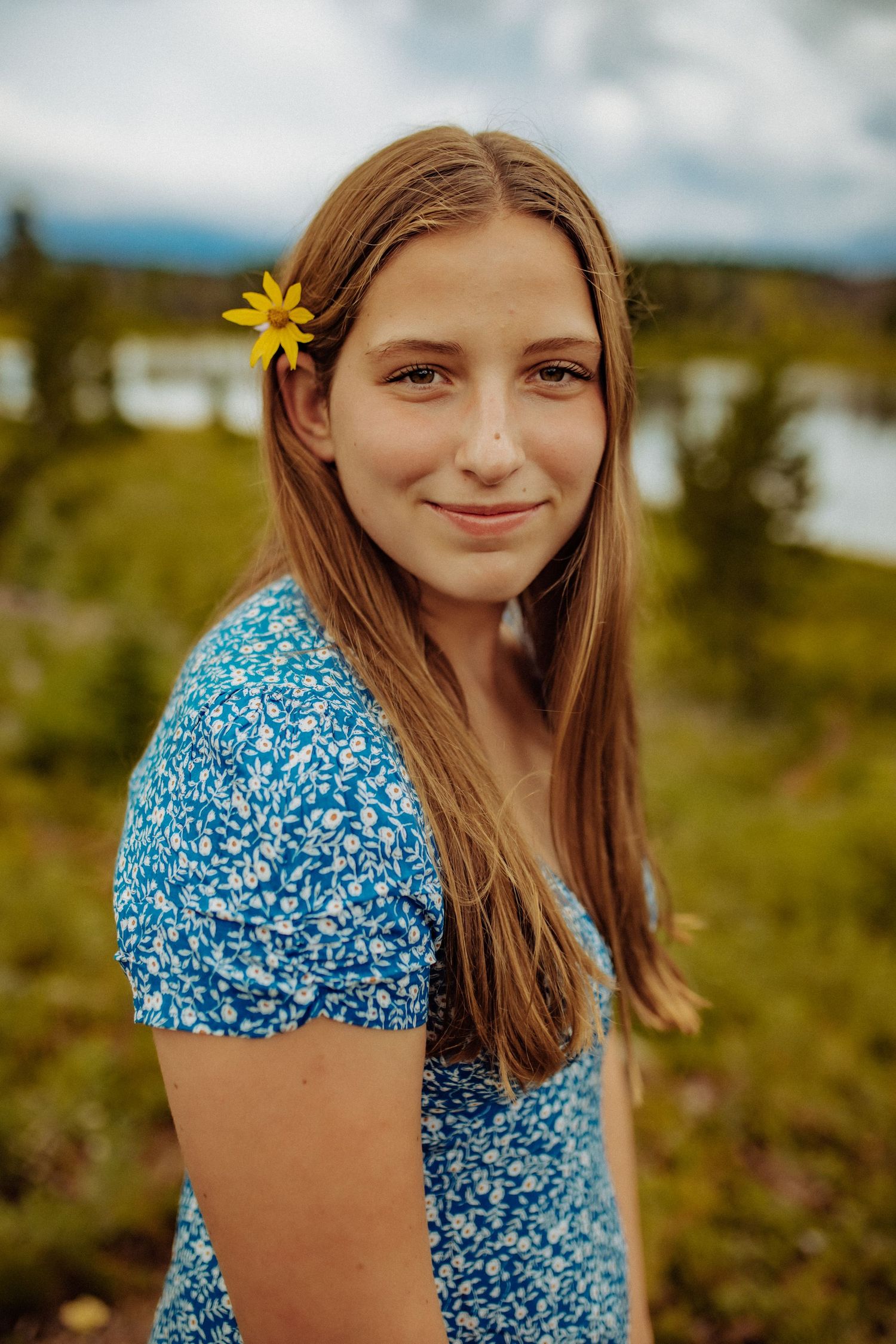 A high school senior girl wearing a blue floral dress stares into the camera lens and has a yellow wildflower tucked behind her ear.