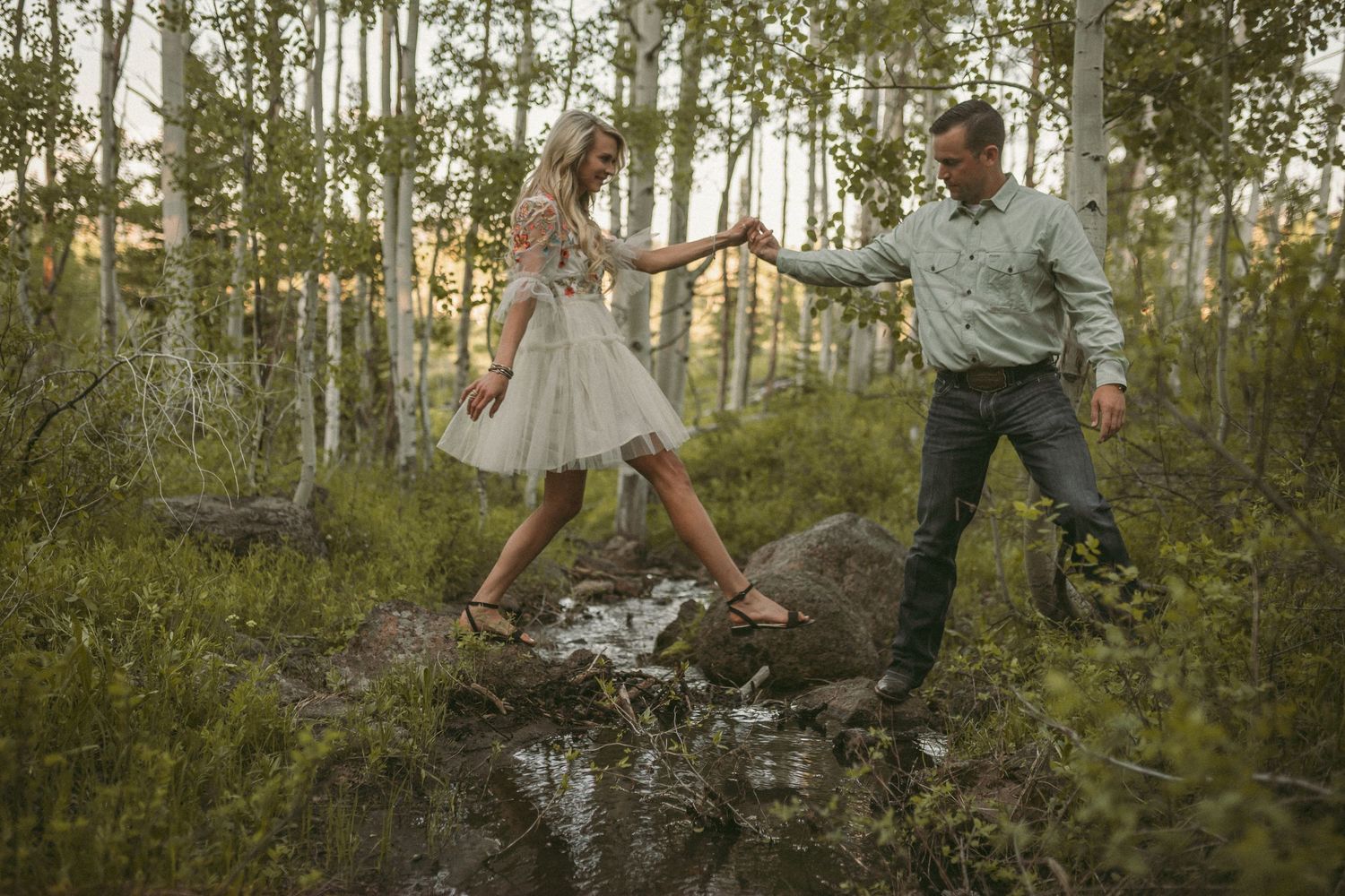 A man helps his girlfriend cross a stream surrounded by aspen trees, photographed by Kristy Baker Photography.