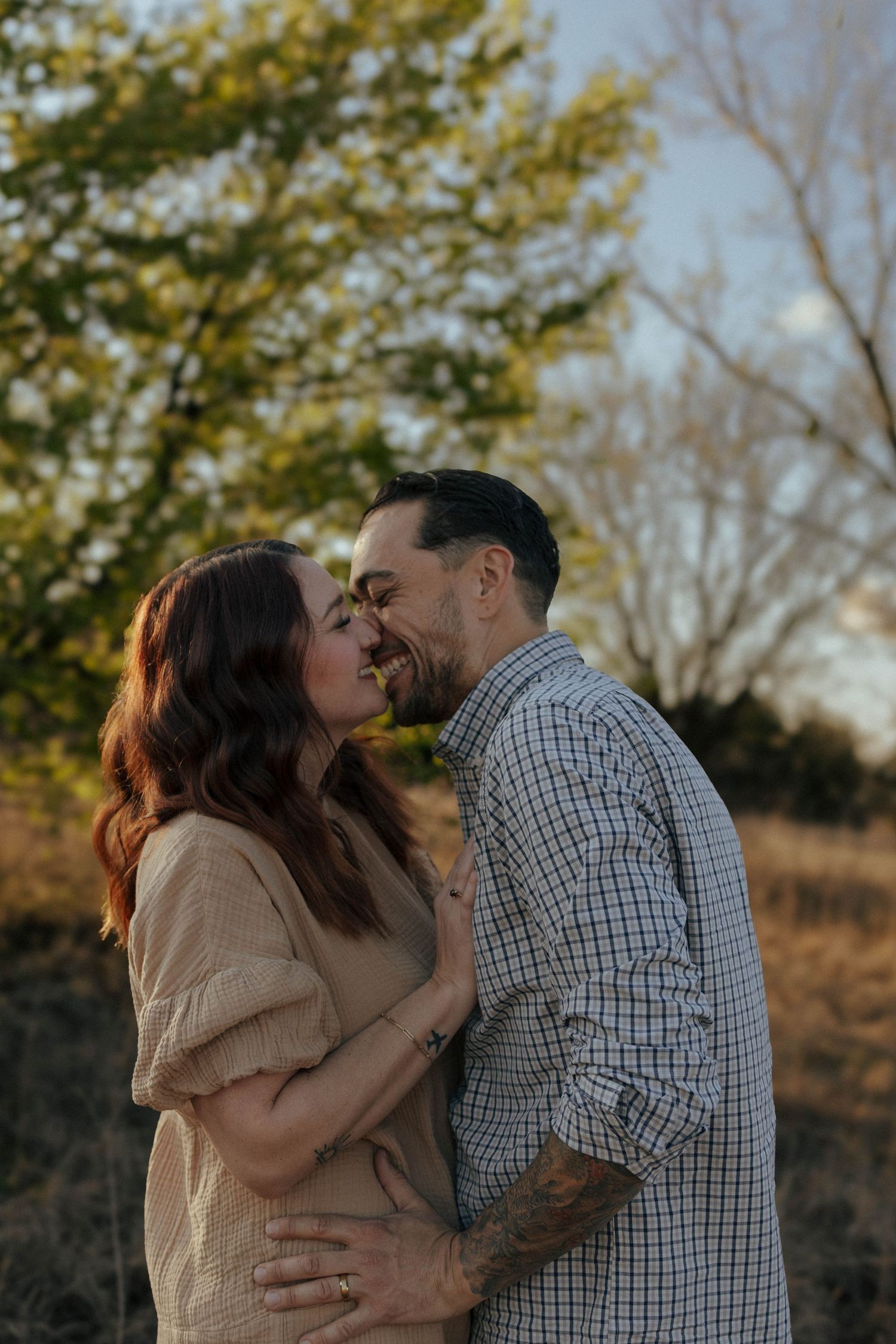 A romantic couple shares tender kisses while embracing outdoors during golden hour in a natural setting.