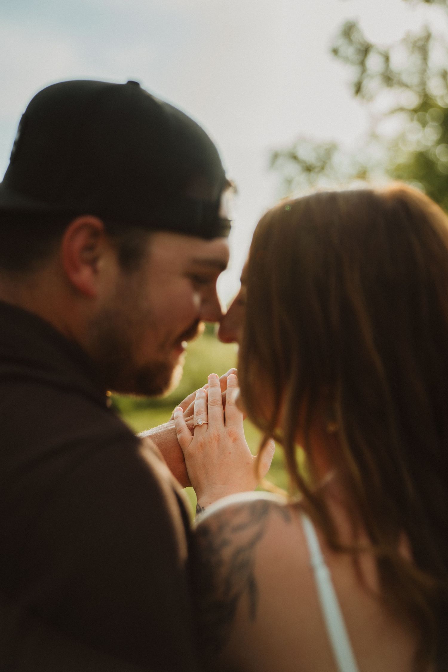 Couple sharing a romantic moment outdoors, man with tattoos and cap holding woman's hand with engagement ring in golden light.