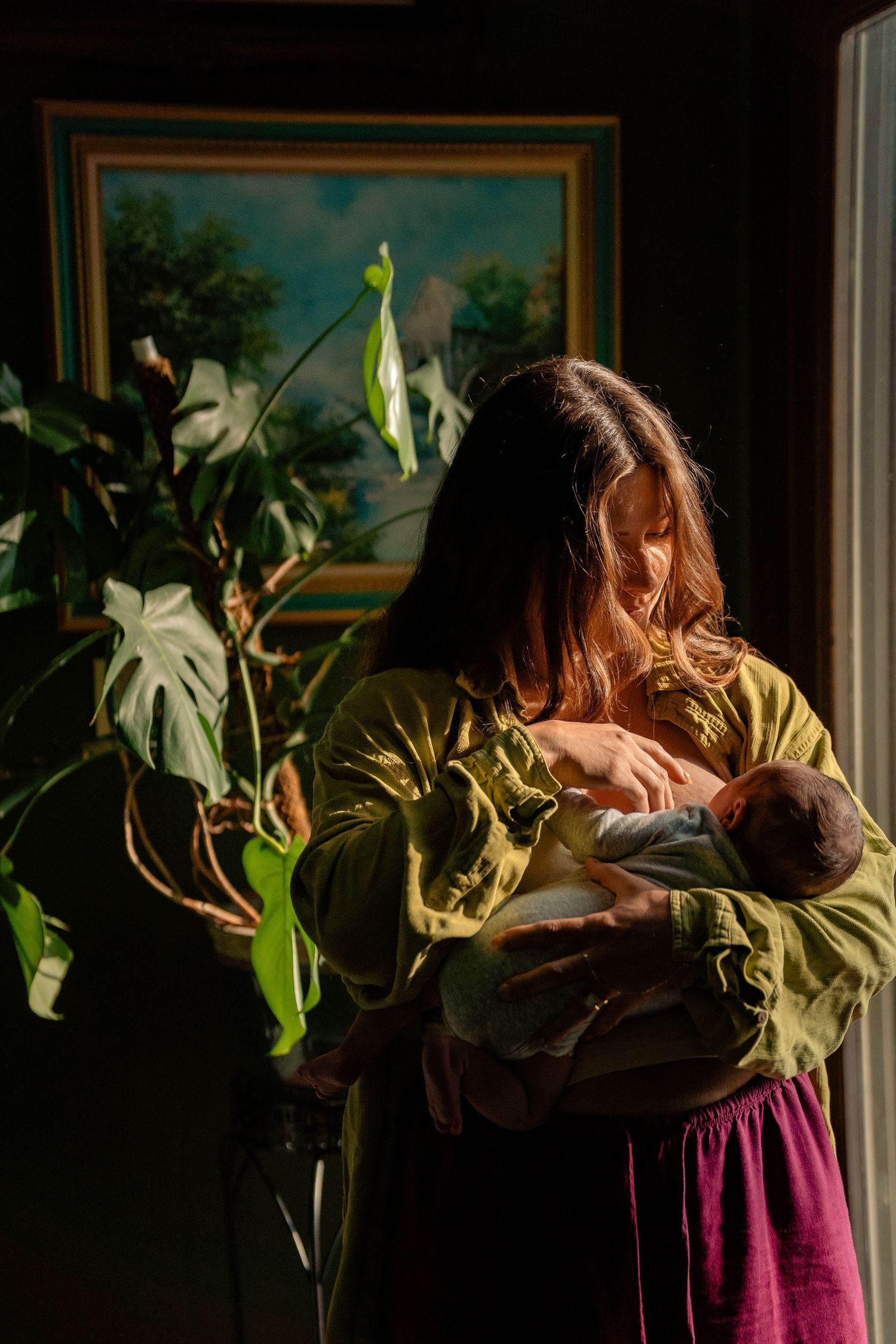 Woman in green blouse holding newborn baby near window with large leafy plant and landscape painting.