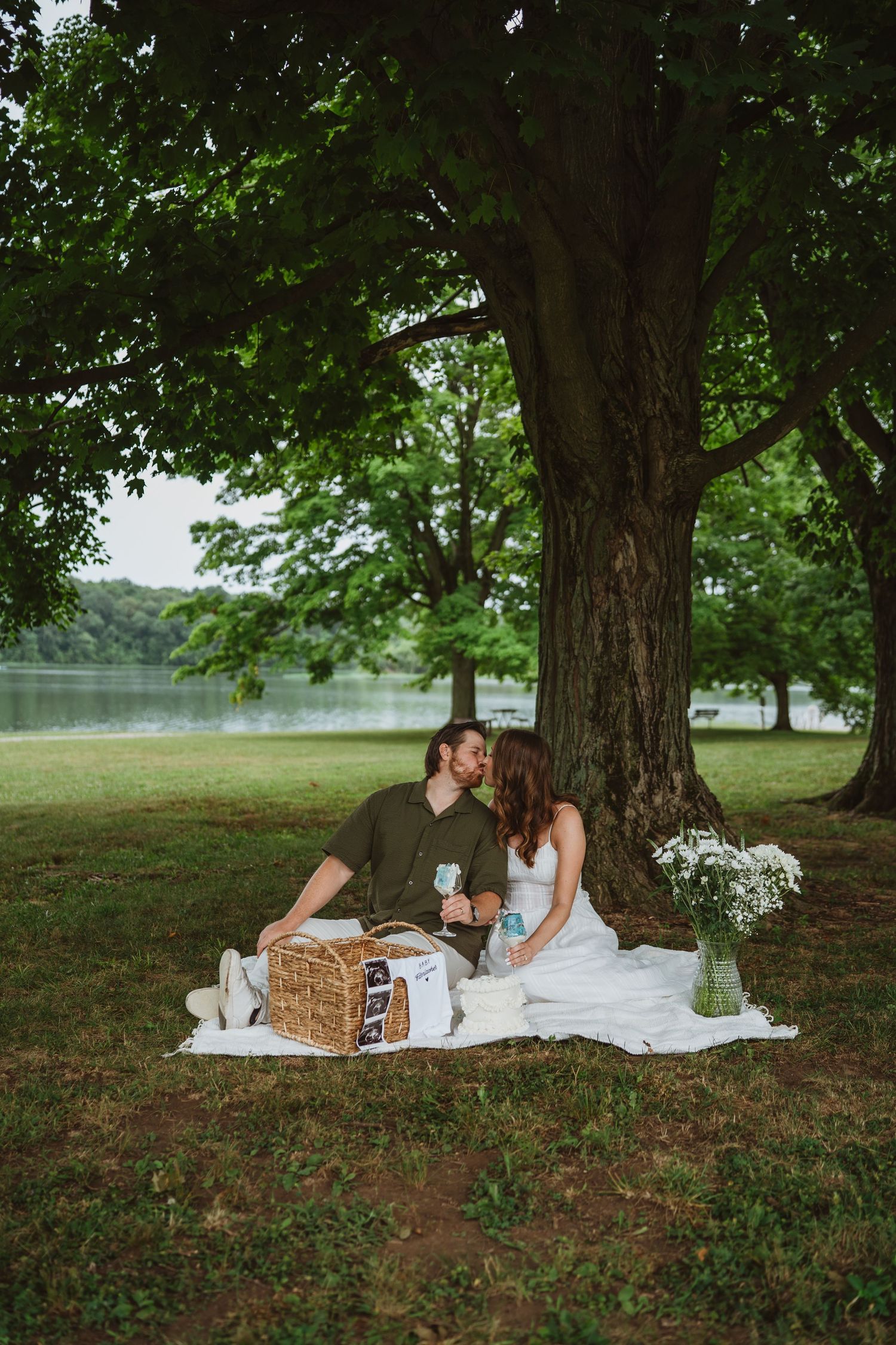 Couple leaning in for a kiss during a lakeside picnic under a large tree, with a wicker basket beside them.