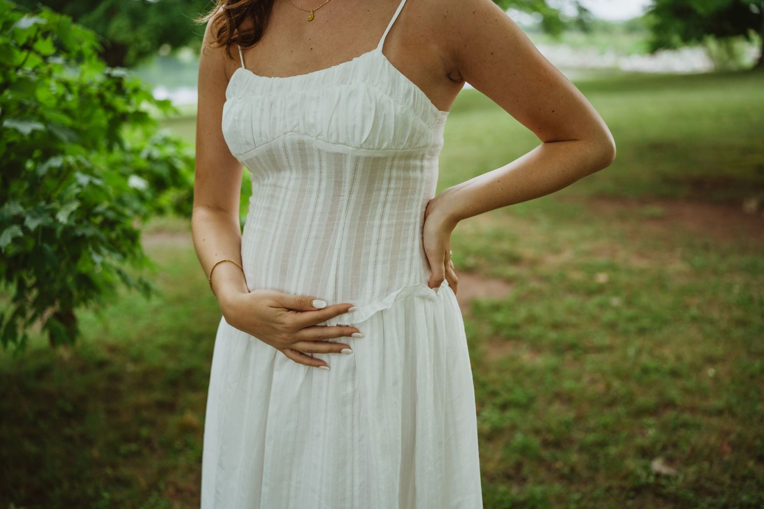 Pregnant woman in a white sundress gently cradling her baby bump in a lush green park.