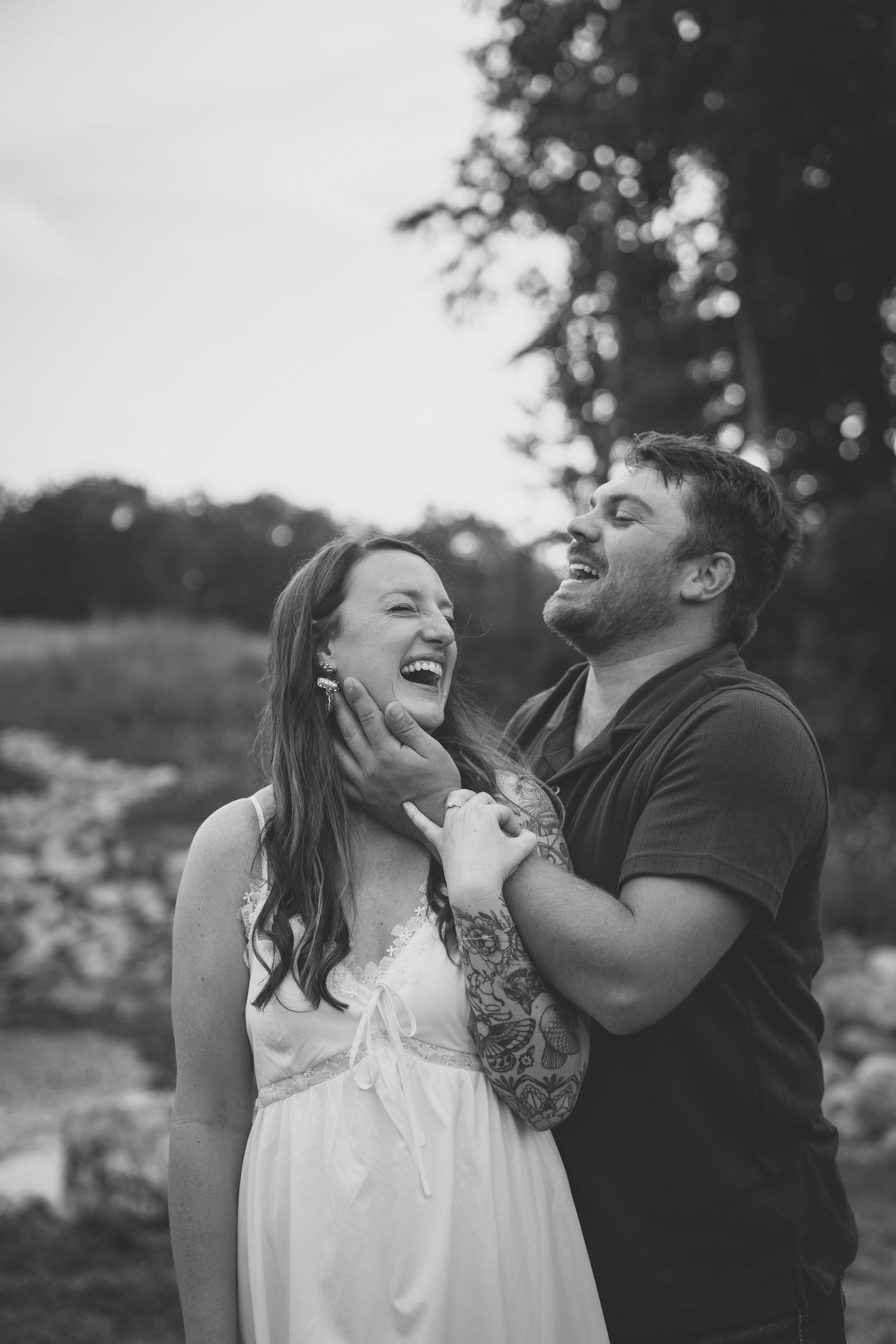 Black and white photo of a laughing couple outdoors, man tenderly holding woman's face, trees in background.