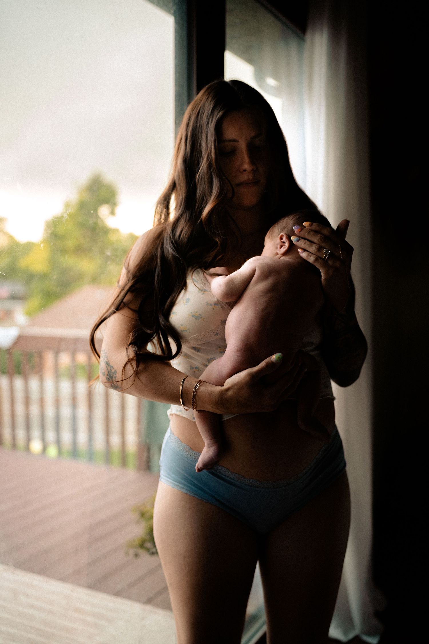 A postpartum mother in lingerie holds her newborn baby near a window at sunset.