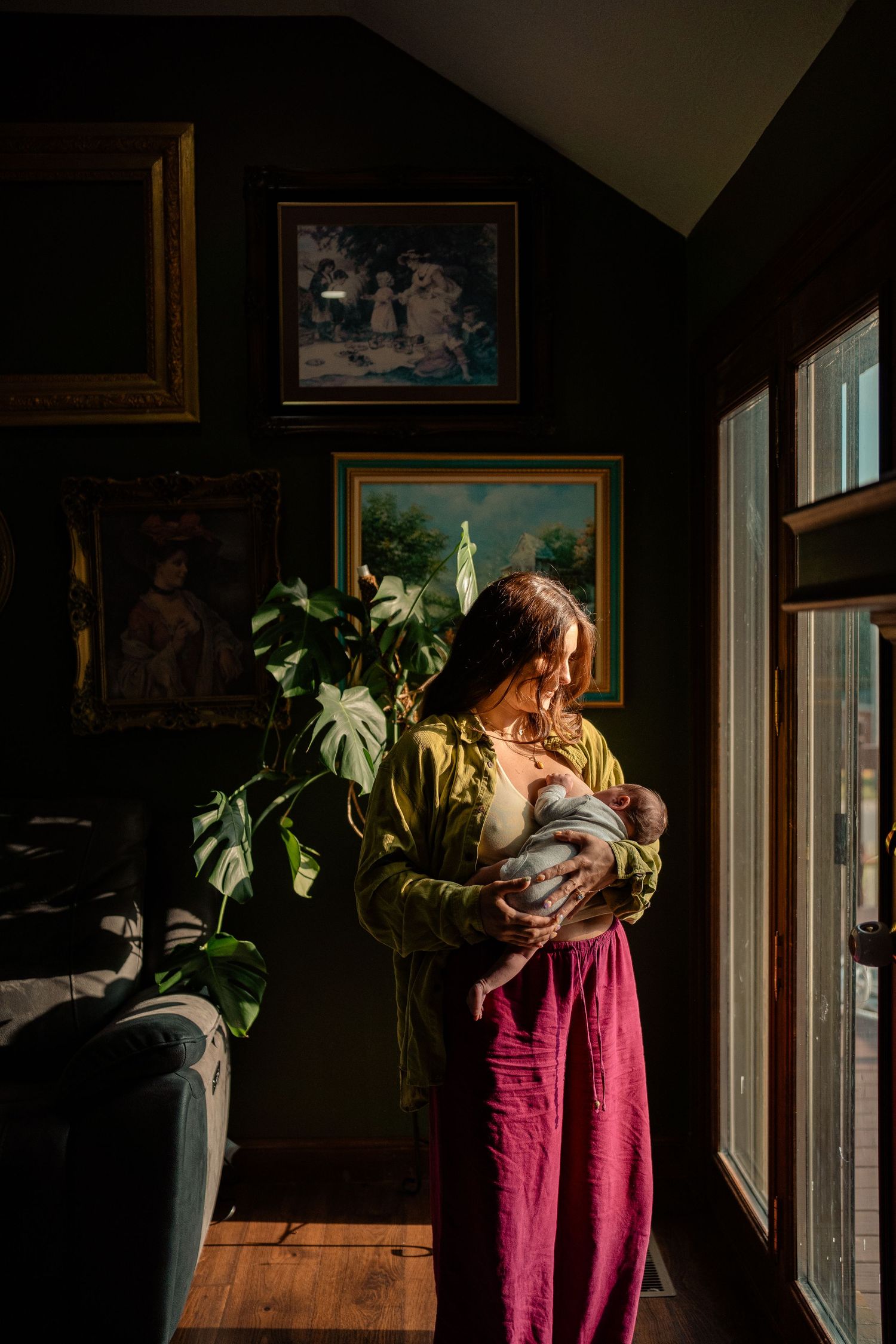 A mother breastfeeds her newborn in a sunlit room adorned with vintage paintings.