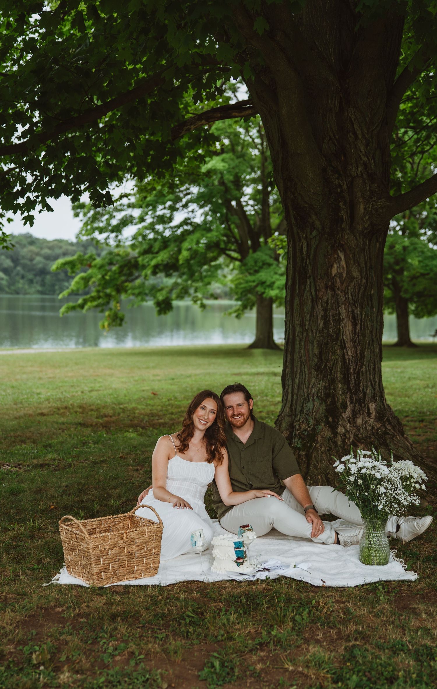 Smiling couple sitting on white blanket under large tree by lake with picnic basket for maternity photos.