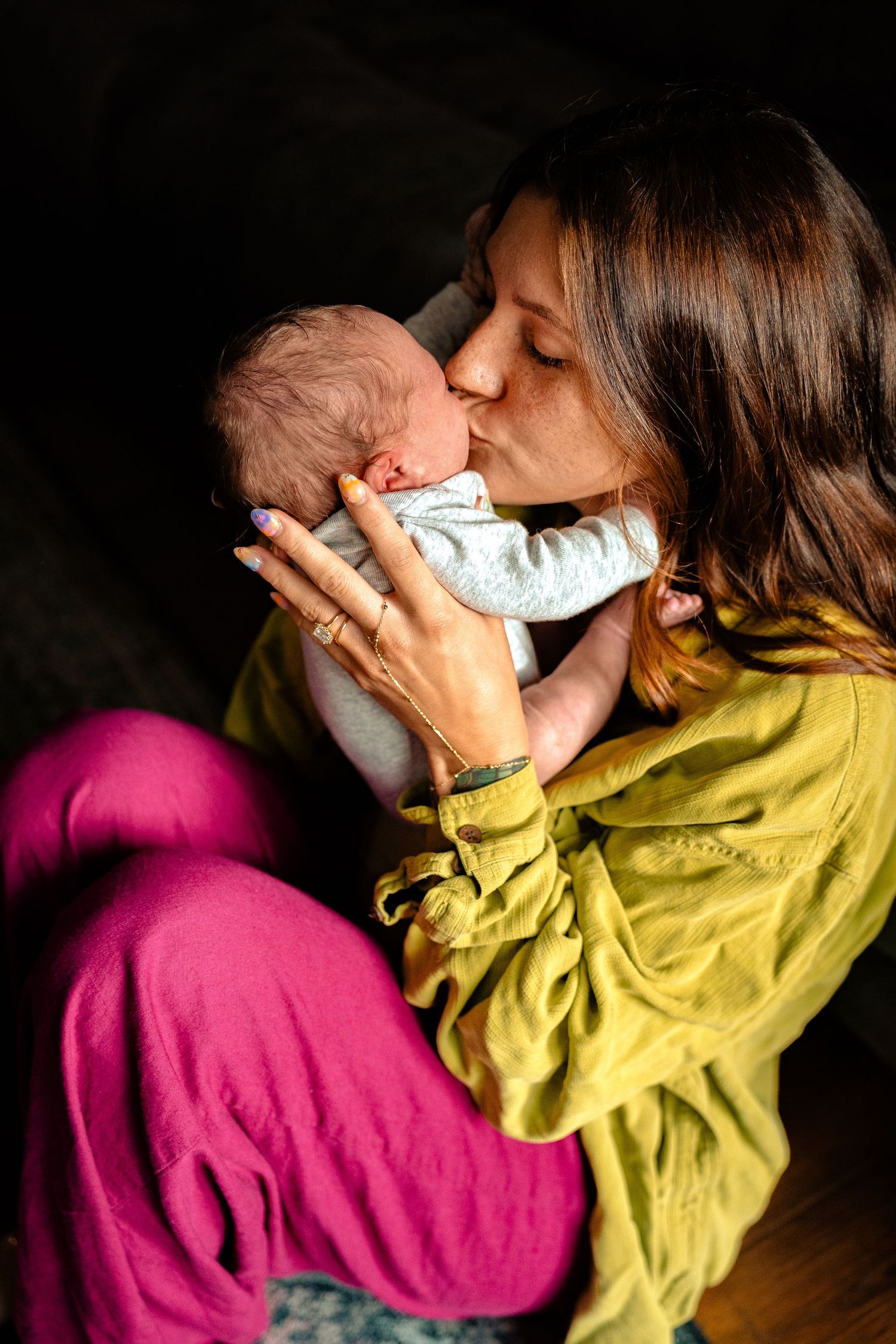 A mother tenderly kisses her newborn baby while cradling it in her arms, wearing a yellow top and pink pants.