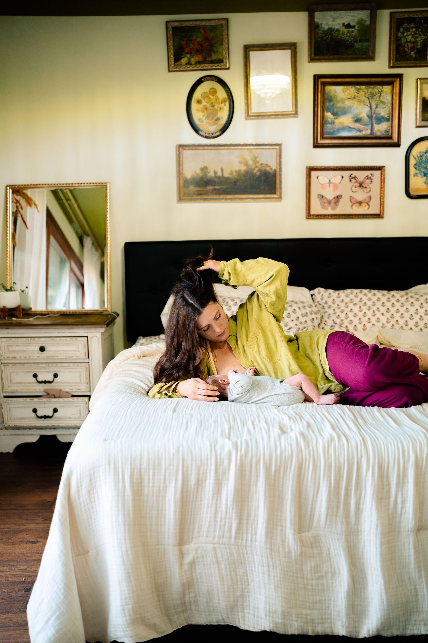 A mother in a yellow top leans over her newborn baby on a white bed.