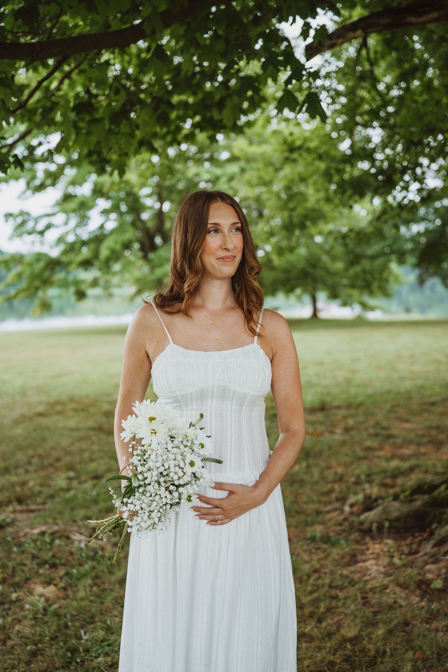 Pregnant woman in white dress holding wildflower bouquet under a tree in a green park.