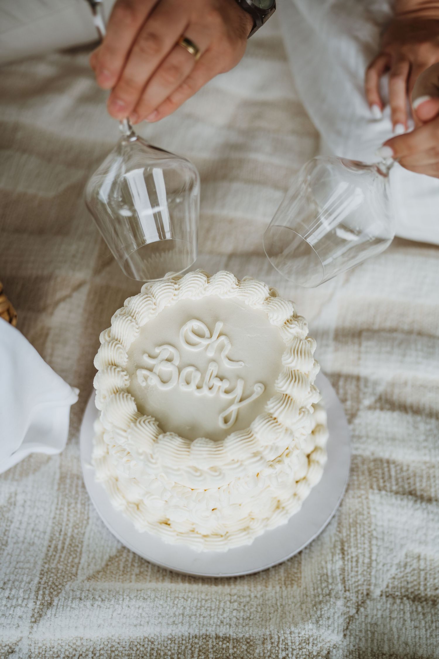 White baby shower cake with Oh Baby inscription, decorated with cream rosettes, as guests clink champagne glasses above.