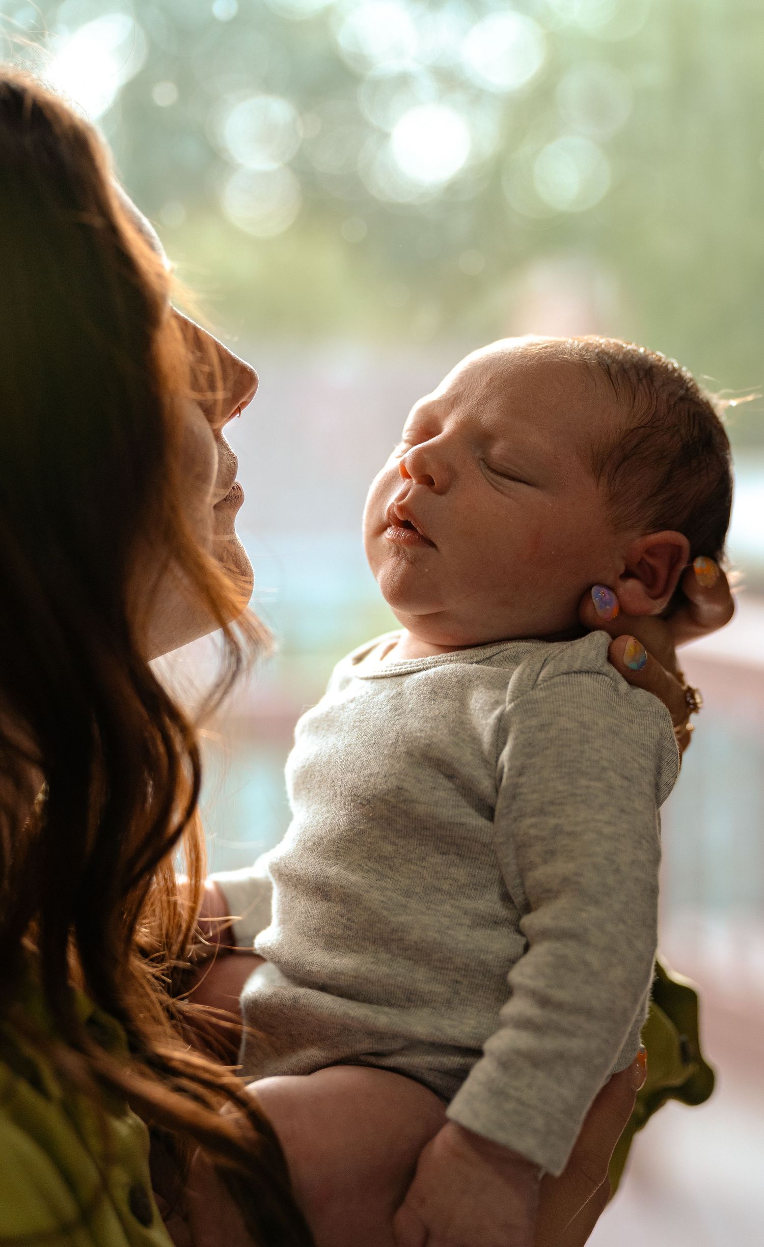 Mother holds sleeping newborn baby in sunlight near bright window, baby wearing gray onesie.