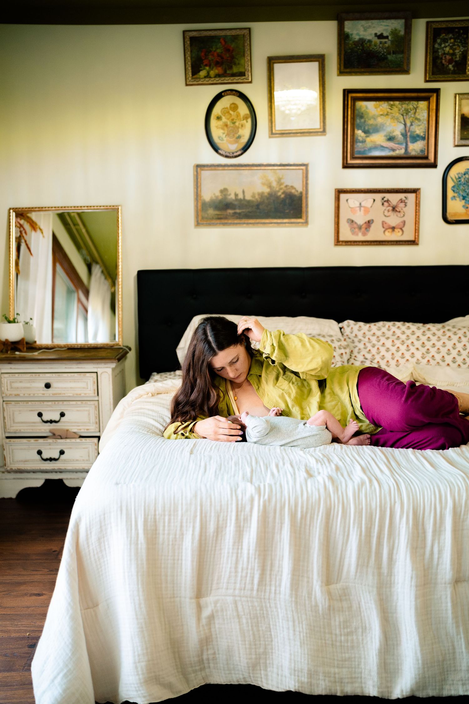 Woman in green robe lying on bed breastfeeding newborn, surrounded by vintage framed artwork on wall.