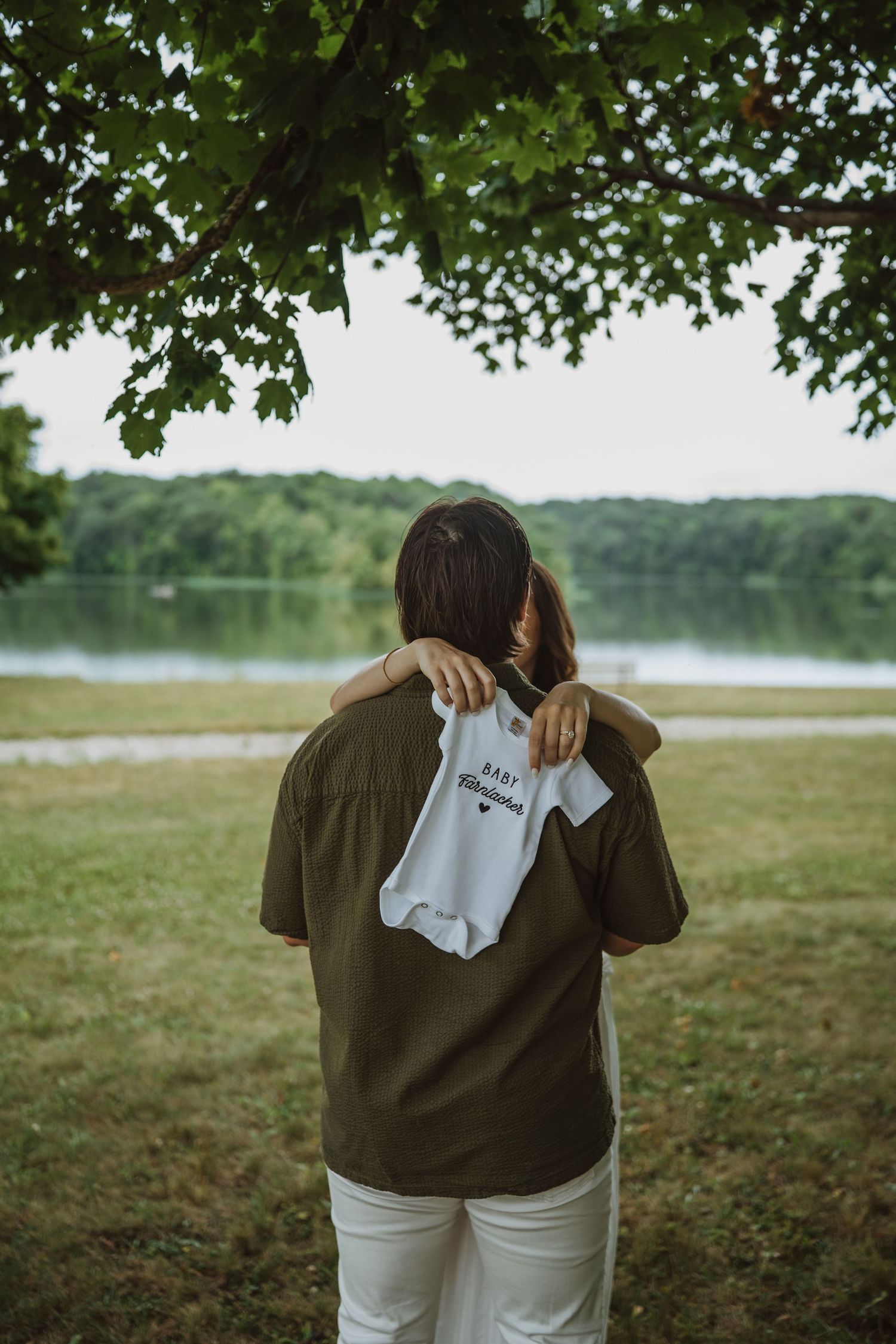 Person holding a white baby onesie behind their back near a lakeside park, announcing pregnancy.