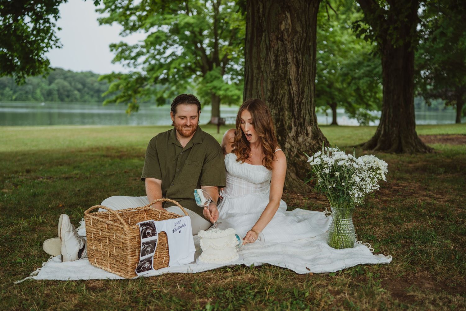 Couple sitting on white blanket under large tree near lake with picnic basket and wildflowers during outdoor maternity photoshoot.